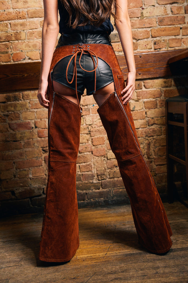 A woman faces a brick wall, wearing INCREDIBLE VELVET DENIM CHAPS with back lacing over black leather shorts. The chaps, open from thigh up, highlight her legs and feature a Western-inspired buckle.