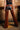 A woman faces a brick wall, wearing INCREDIBLE VELVET DENIM CHAPS with back lacing over black leather shorts. The chaps, open from thigh up, highlight her legs and feature a Western-inspired buckle.