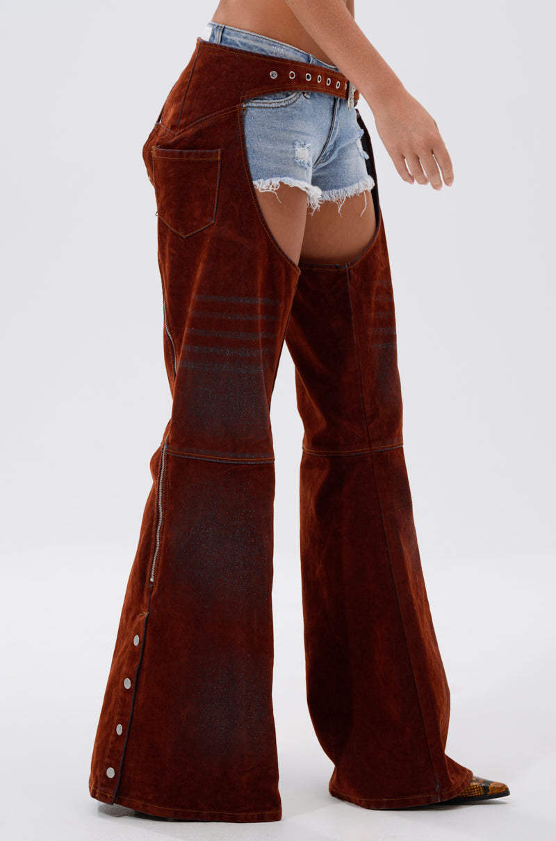 A person stands against a plain background, channeling cowgirl couture in INCREDIBLE VELVET DENIM CHAPS featuring open sides, flared legs, and distressed denim shorts underneath.