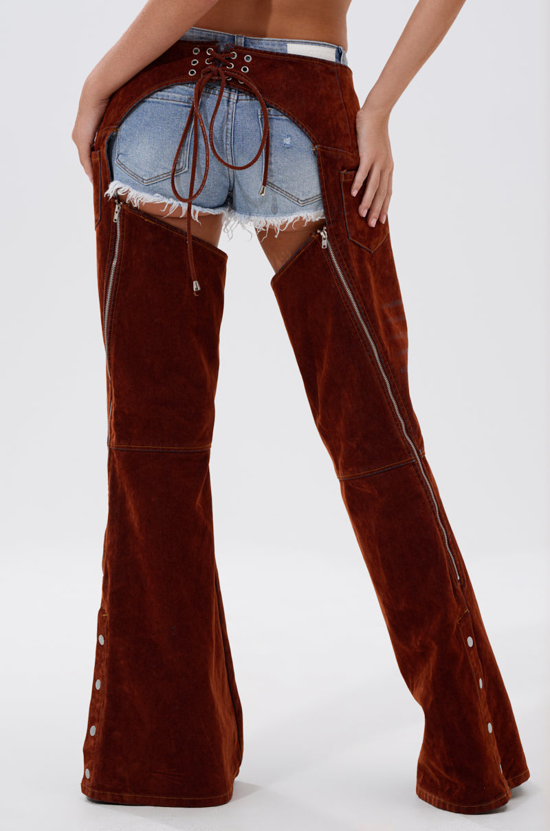 A person, shown from the back, wears INCREDIBLE VELVET DENIM CHAPS over frayed shorts. These chaps feature lace-up waist detailing, side zippers, and wide legs for a statement cowgirl couture look. The upper body is not visible.
