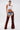 A woman stands confidently against a plain white background, wearing a white cropped tank, layered necklaces, distressed denim shorts with a belt, and INCREDIBLE VELVET DENIM CHAPS for a modern cowgirl look.