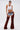 A woman in oversized INCREDIBLE VELVET DENIM CHAPS, short denim shorts, a white crop top, layered necklaces, clear glasses, and a studded belt exudes cowgirl couture against a plain background.