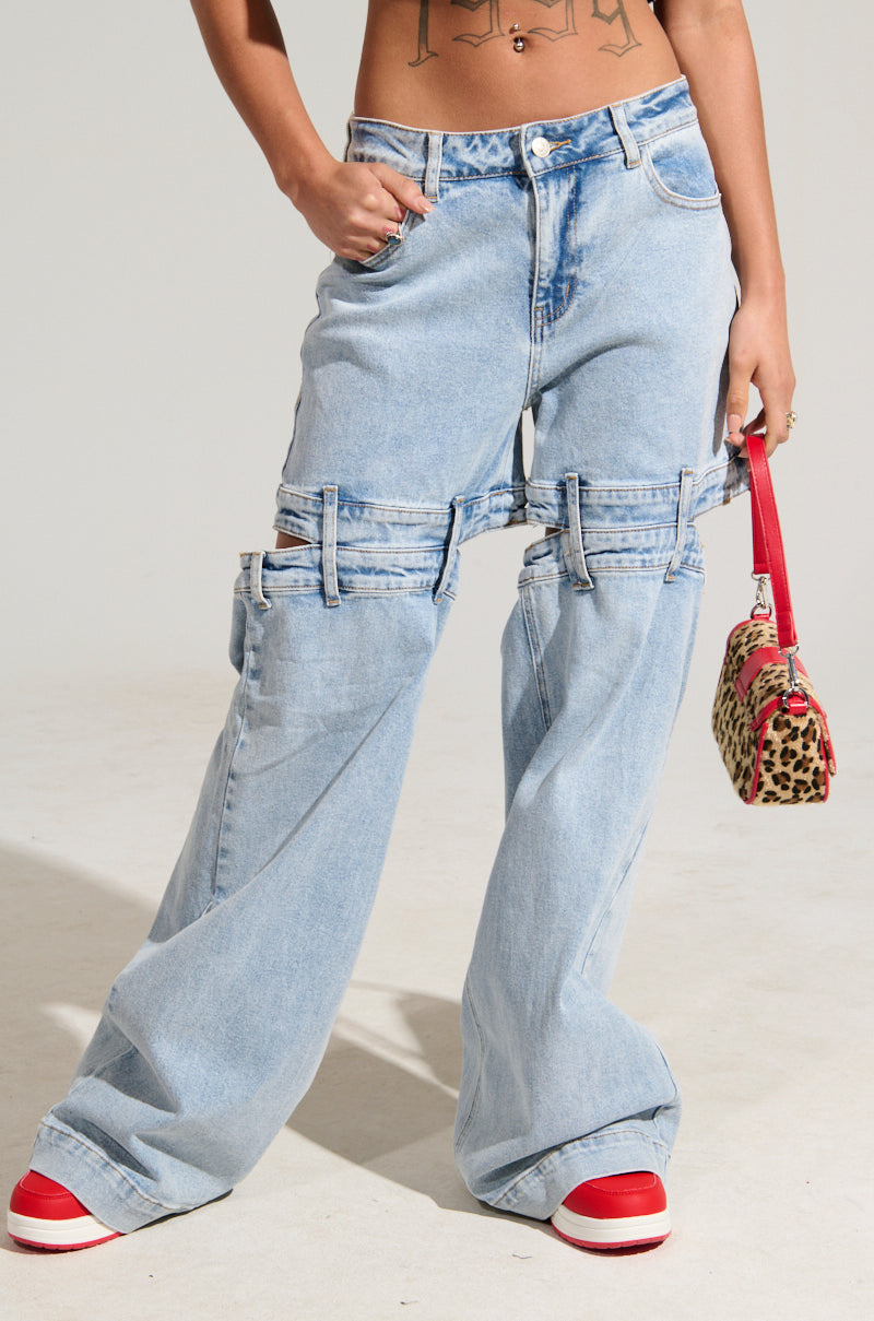A person is wearing the LET LOOSE DENIM PANT in light blue, characterized by a high-rise waist and wide leg silhouette with cut-out details. They are also sporting red-and-white sneakers and carrying a leopard print handbag with a red strap, showcasing a tattoo on their midriff.