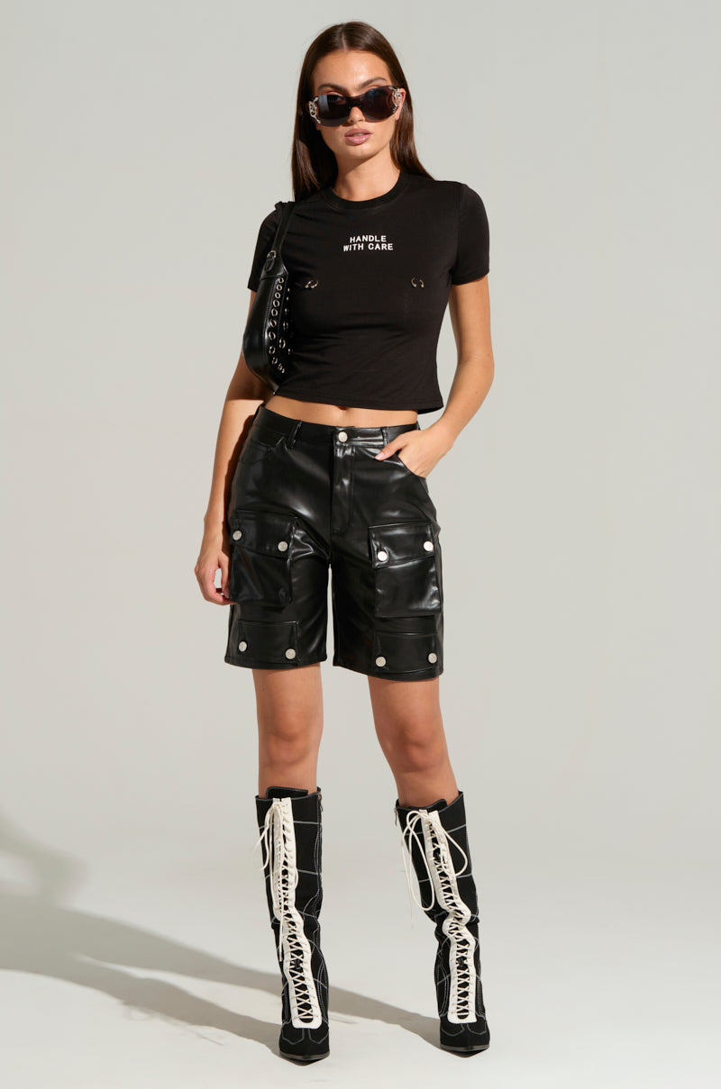 A woman stands confidently wearing a black crop top, the LERAY FAUX LEATHER BERMUDA SHORTS IN BLACK with silver accents, tall black lace-up boots, and sunglasses. She has a black purse draped over her shoulder and her hands resting on her hips. The background is plain and light-colored.