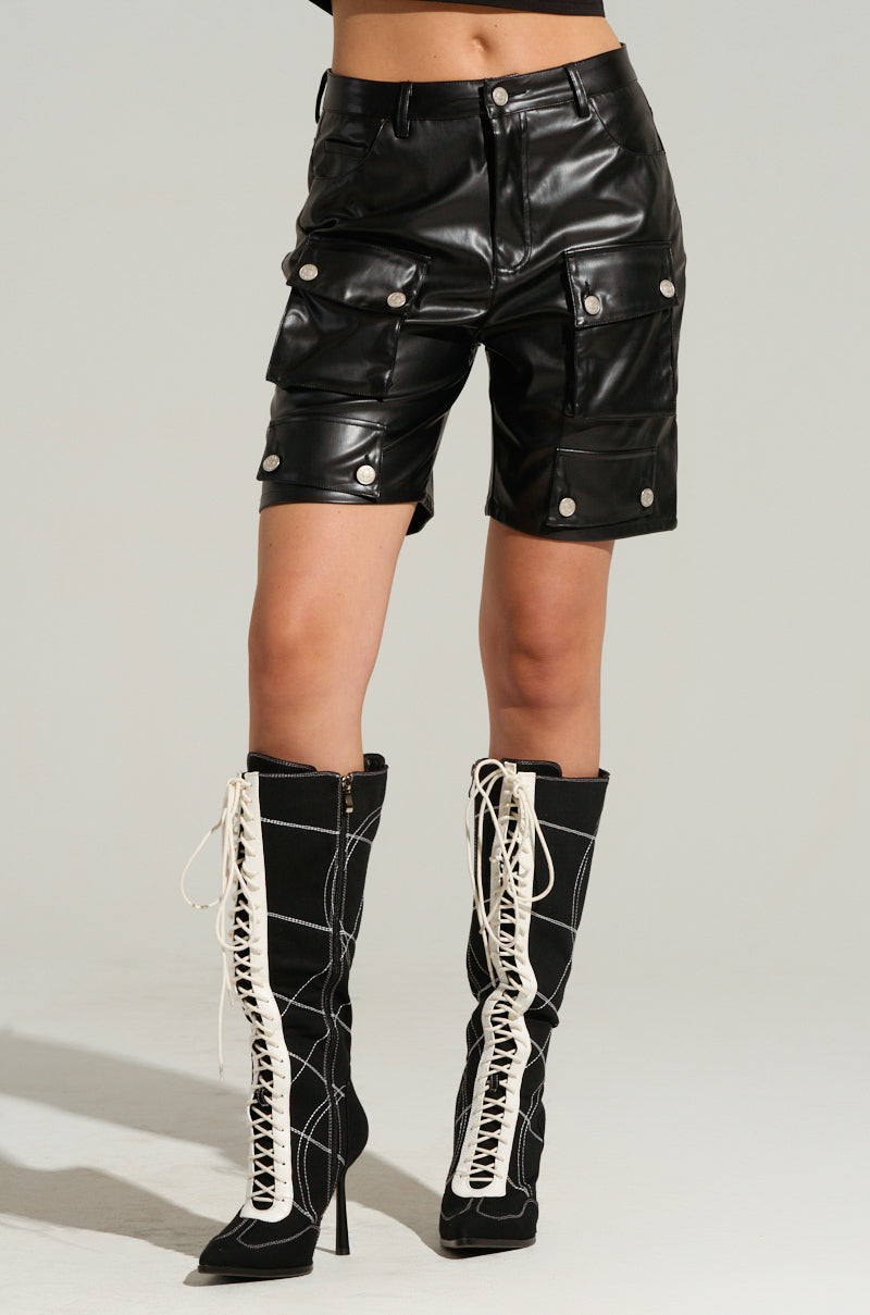 A person is wearing black knee-high boots with intricate white laces and zippers. They are also sporting the LERAY FAUX LEATHER BERMUDA SHORTS IN BLACK, which feature large pockets and silver buttons. The background is neutral and plain.