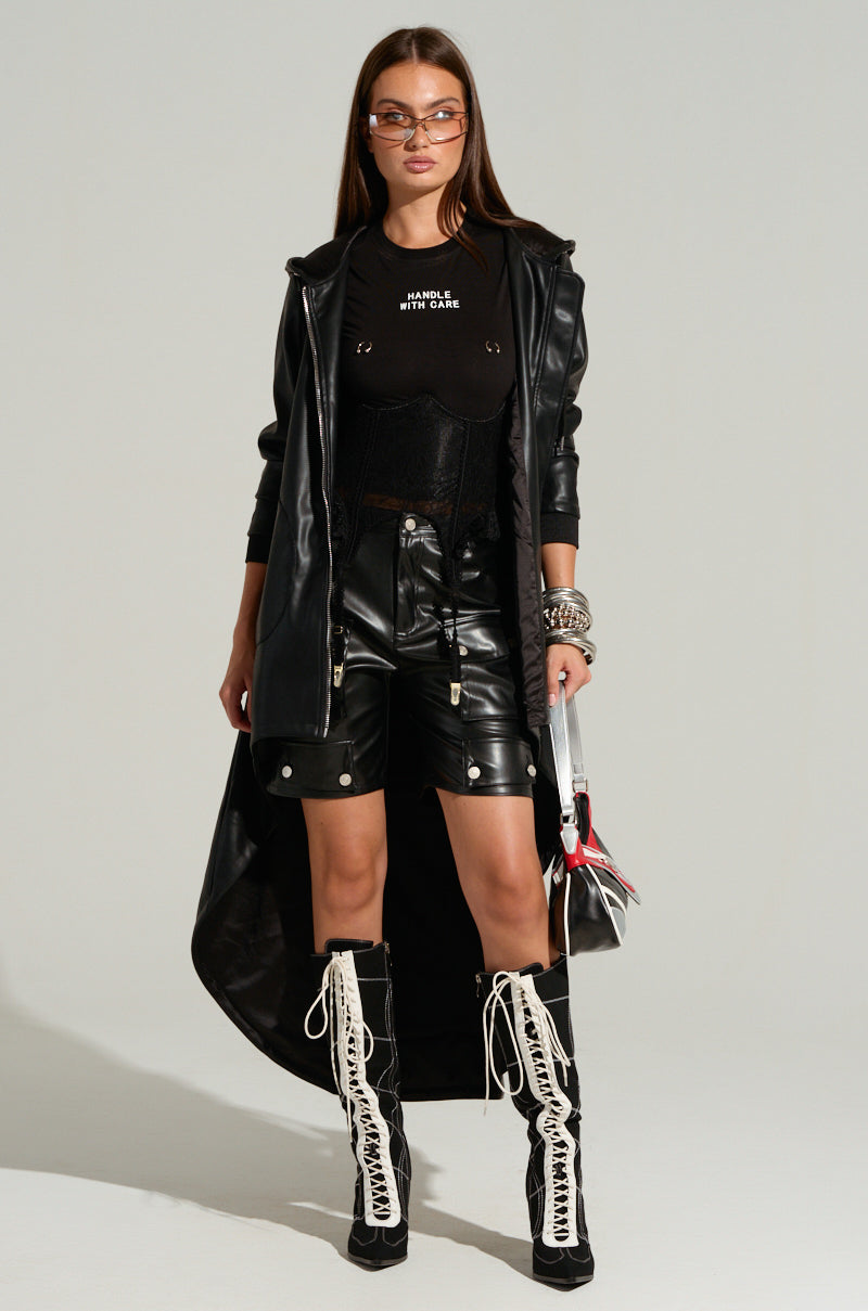 A person stands confidently wearing a black leather jacket with a long train, black shirt, LERAY FAUX LEATHER BERMUDA SHORTS IN BLACK, and black boots with white lace-up detailing. They sport sunglasses and assorted silver bracelets while holding a small bag in their right hand.
