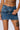 A person's midsection and upper legs wearing the super stretchy JUST WANNA ROCK DENIM MINI SKIRT, featuring a frayed waistline with metal eyelets, a grommet belt, and chain detail. Their lower torso has visible tattoos, including "1999" across the abdomen and a stylized face on the thigh.