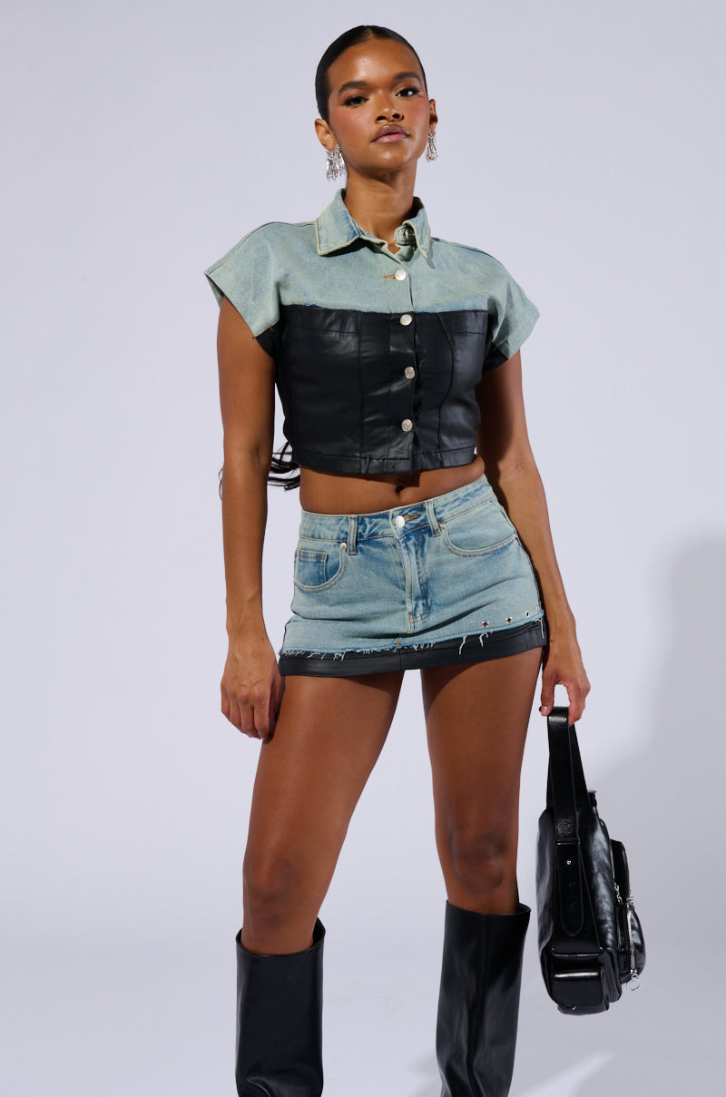 A woman poses confidently in a stylish outfit, featuring a light blue and black color-blocked crop top with buttons, the YOU GIVE ME DENIM MINI SKIRT with distressed detailing, and black knee-high boots. She holds a black bag and has her hair pulled back against a plain background.