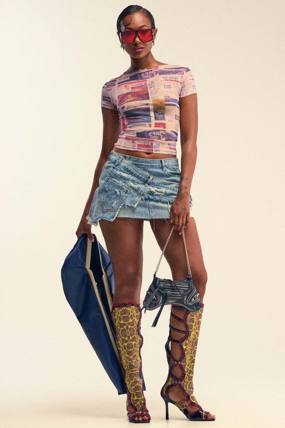 A woman poses against a plain background wearing red sunglasses, a patterned fitted top, knee-high yellow lace-up boots, and the DUMP HIM LOW RISE DENIM MINI SKIRT. She holds a large blue bag and a small purse.