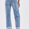 A person wearing IN THE MOOD DENIM PANT with cuffed hems and heeled sandals stands facing away, highlighting the straight-leg fit, two back pockets, and a visible brand label on the waistband.