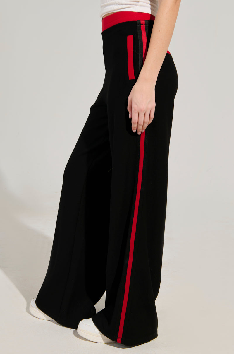 A person wearing the VARSITY LEAGUE STRIPED WOVEN ELASTIC TROUSER IN BLACK RED, featuring sporty side seam stripes, paired with a white top and white sneakers, is standing against a plain background. These trousers add a bold accent to the outfit.