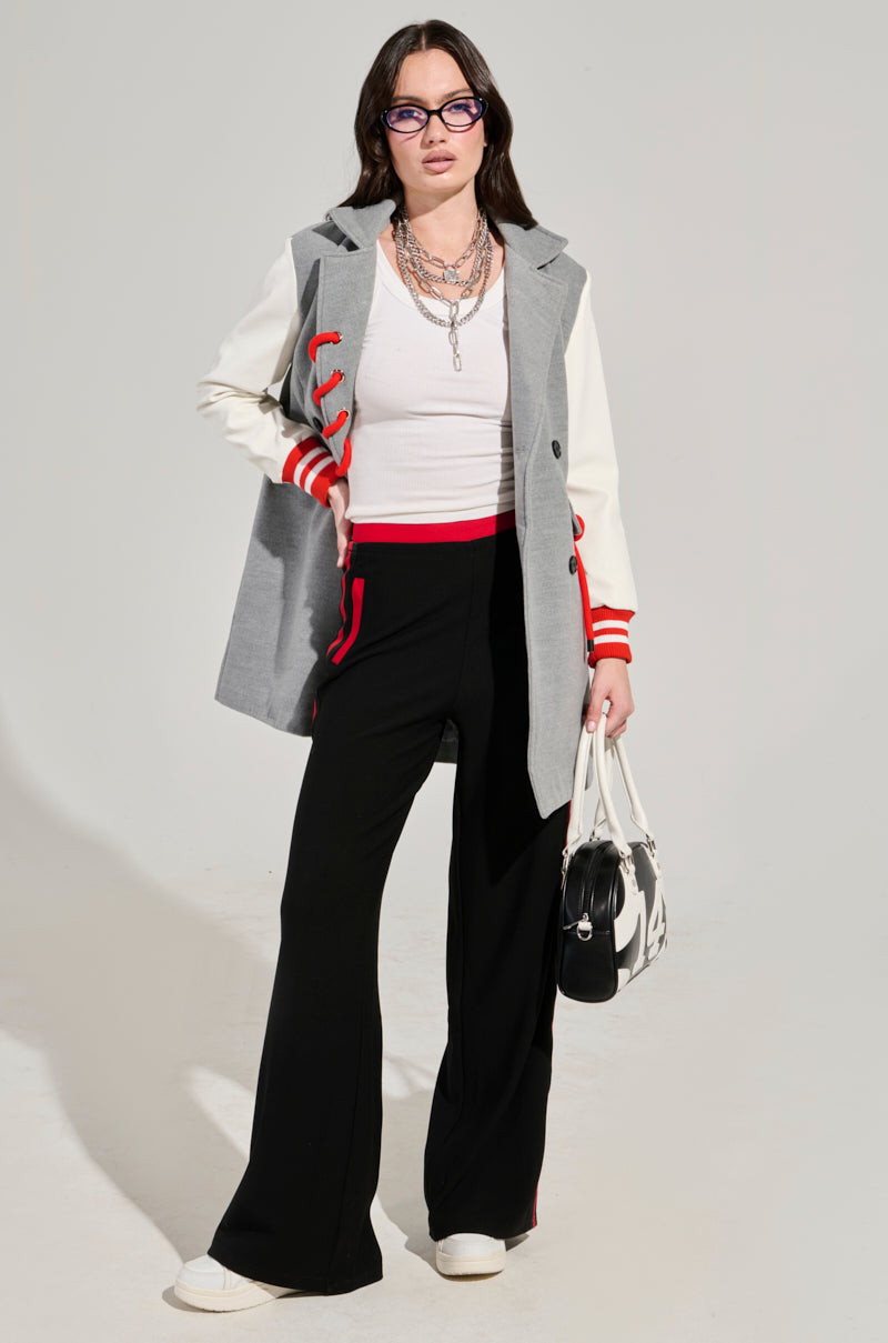 A woman with long dark hair wears glasses and a gray coat adorned with red lace-up details over a white shirt. She pairs her outfit with the Varsity League Striped Woven Elastic Trouser in Black Red, showcasing high-rise wide-leg pants highlighted by red accents. Her ensemble is finished off with white sneakers and a black and white bag, as she confidently poses against a plain background.