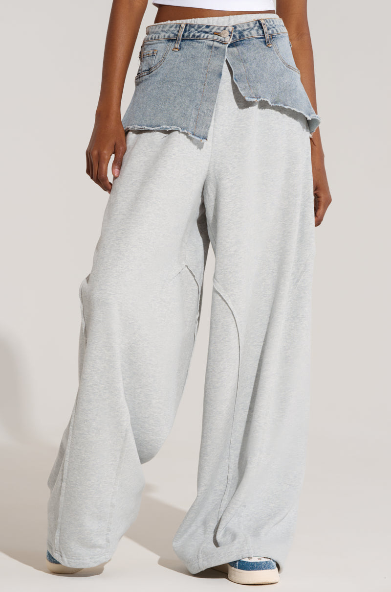 A person is wearing light grey DYLAN DENIM BELTED SWEATPANTS with a wide leg silhouette, paired with frayed, cut-off denim shorts layered over the waistband. The look is completed with white sneakers featuring blue accents. The background is a plain light grey color.