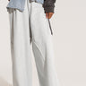 A person is wearing the DYLAN DENIM BELTED SWEATPANT, which features loose-fitting gray sweatpants paired with a unique worn, frayed denim overlay that resembles the upper part of a pair of jeans without the legs. The outfit boasts a wide leg silhouette and high rise design, complemented by a dark jacket. The image is cropped from the waist down.