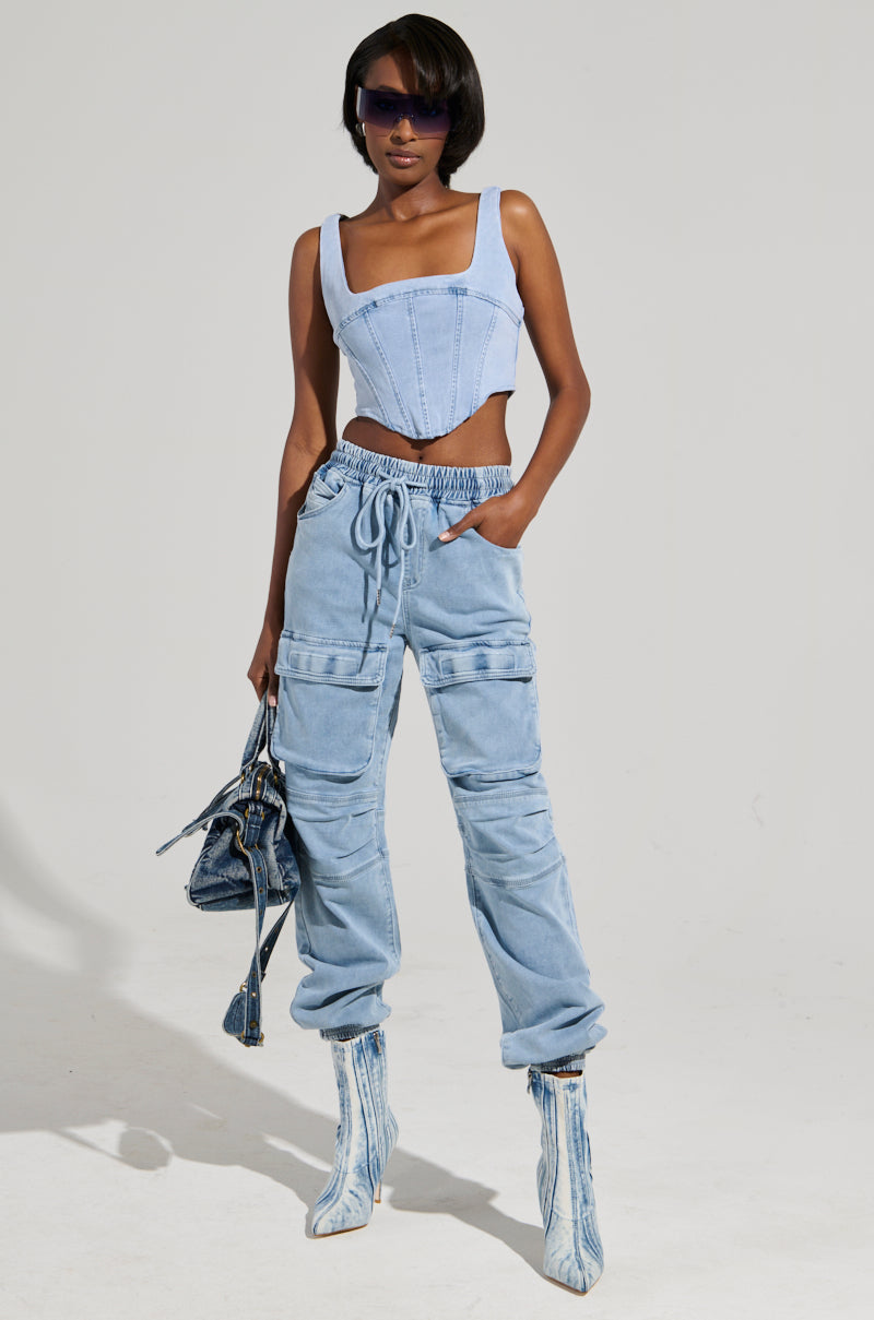 A person stands confidently wearing a light blue corset top and the STAY COOL DENIM JOGGER, featuring a relaxed-fit and high-rise design. They accessorize with sunglasses, a stylish denim handbag, and patterned ankle boots against a simple light gray background.
