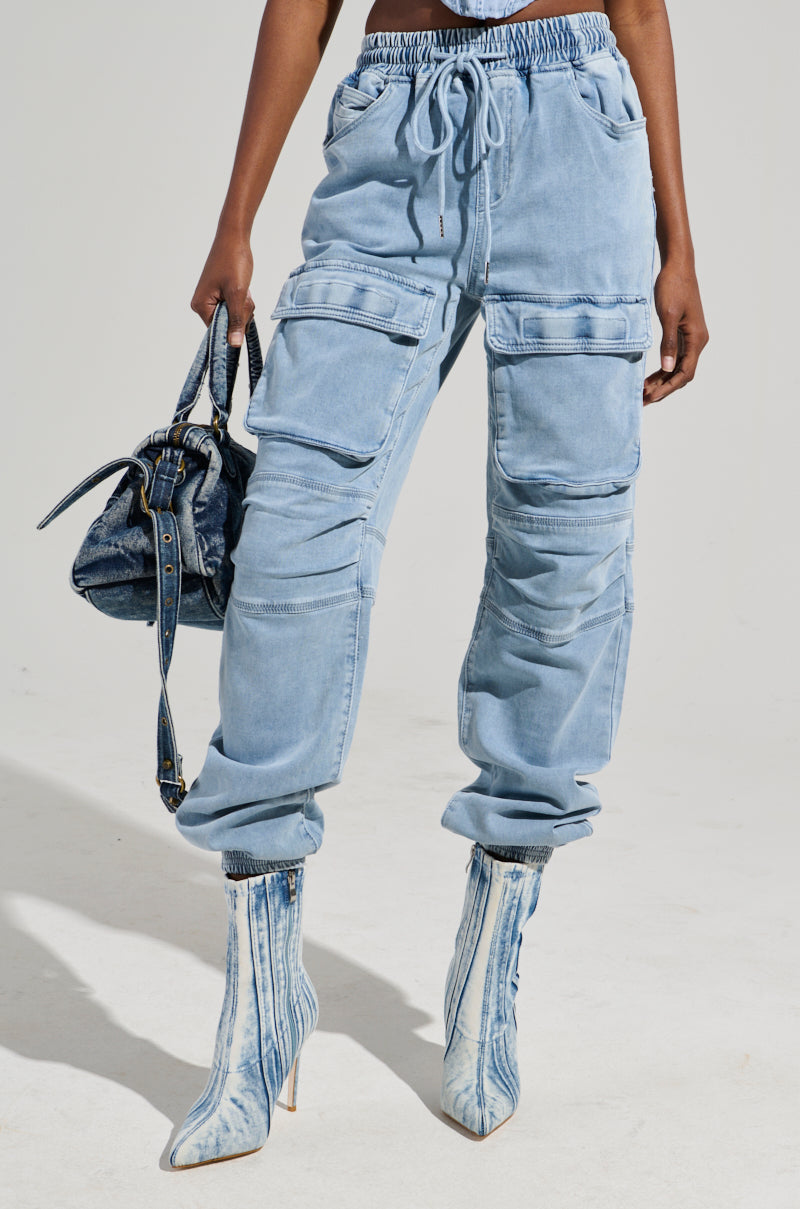 A person wearing STAY COOL DENIM JOGGER pants in a relaxed fit, featuring light blue denim with multiple pockets and a drawstring waistband. They pair it with high-heeled, faded denim boots and carry a dark blue denim handbag against a plain, neutral background.