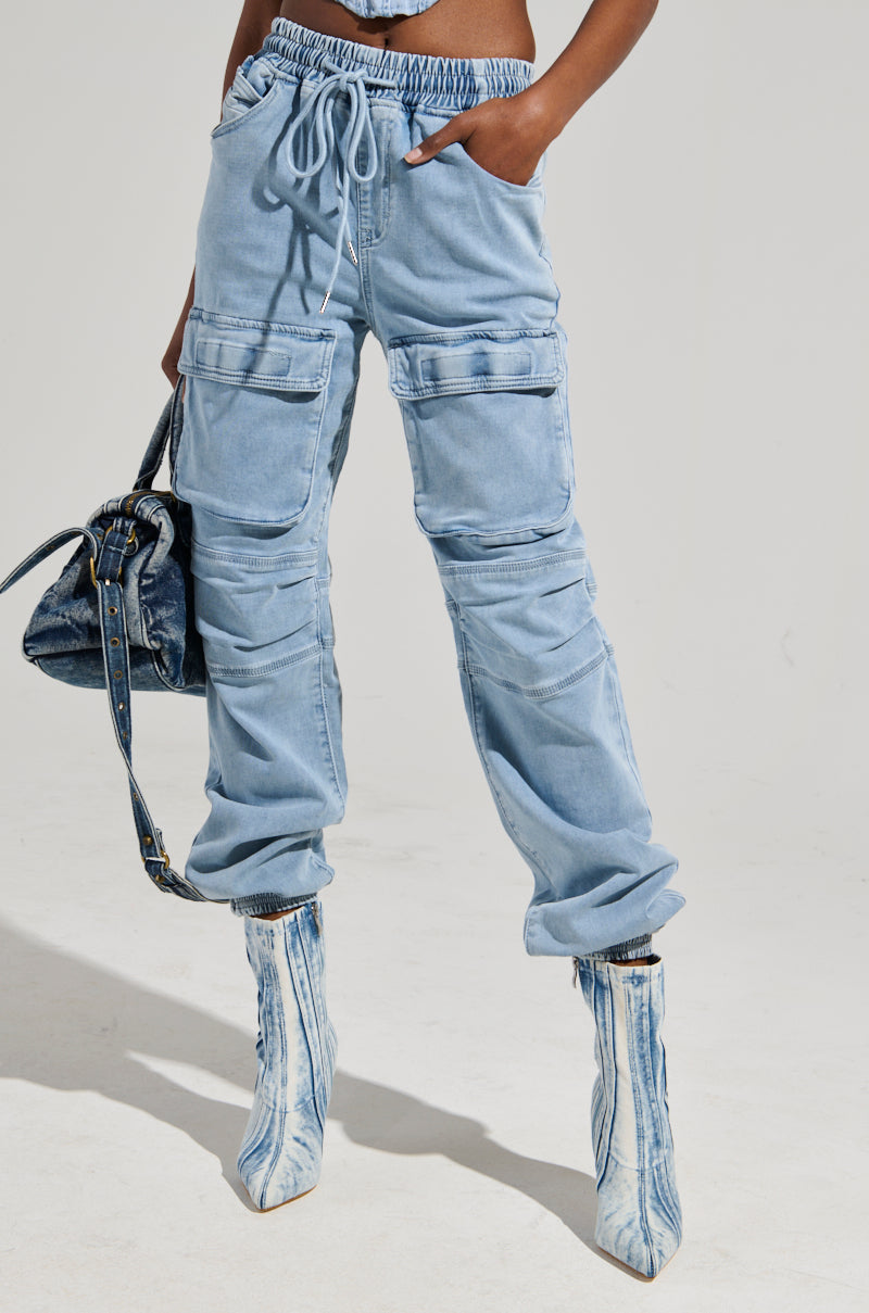 A person wearing the STAY COOL DENIM JOGGER in relaxed-fit light blue with multiple pockets and a drawstring waist, paired with matching denim boots. They carry a dark blue denim bag against a plain white background.