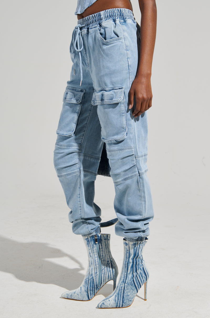 A person wearing the STAY COOL DENIM JOGGER, featuring a light blue hue, high-rise design, drawstring waist, and multiple pockets. Paired with high-heeled boots in a matching denim pattern, this relaxed fit ensemble stands out against the plain background, enhancing the focus on the outfit.
