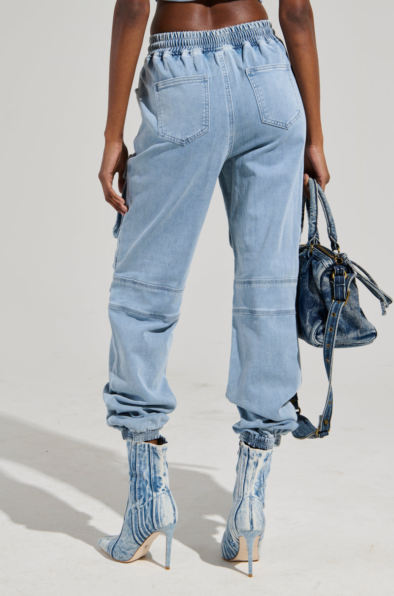 A person dressed in STAY COOL DENIM JOGGERs in a relaxed-fit light blue style, paired with high-heeled boots featuring a tie-dye pattern, while holding a blue handbag. The image captures the lower half of the individual against a plain background.