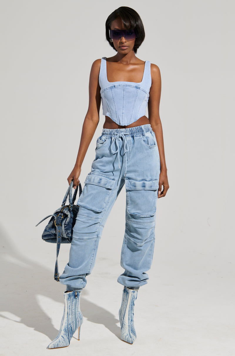 A person in a denim corset top and STAY COOL DENIM JOGGER pants poses confidently with denim boots. They hold a denim handbag and wear sunglasses.