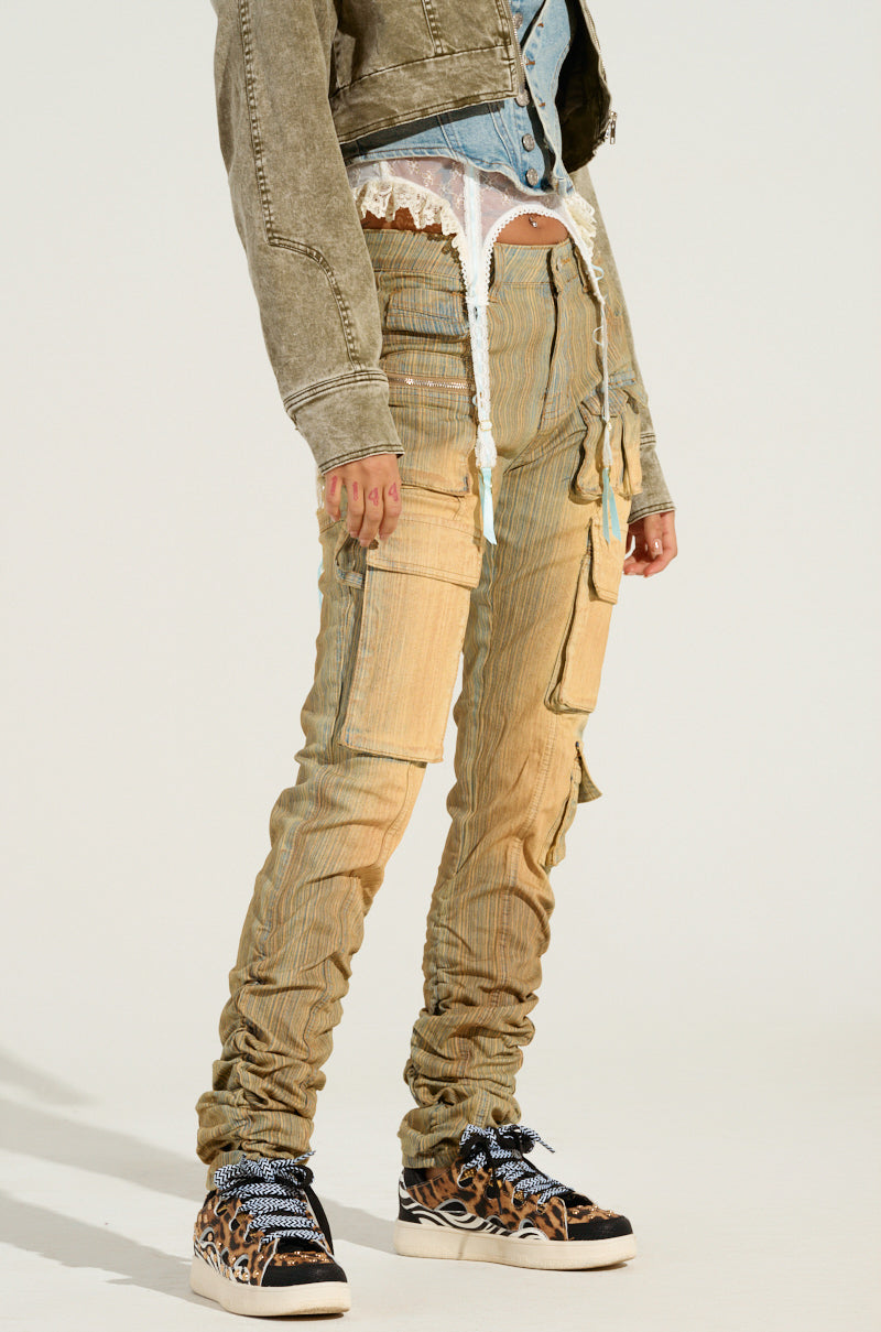 A person showcases an ensemble featuring a green denim jacket over a denim shirt and white lace bralette, styled with the NATALIE OMBRE EFFECT SKINNY CARGO PANT in beige and complemented by black sneakers adorned with orange tiger stripes. The person casually rests their hands in their pockets.