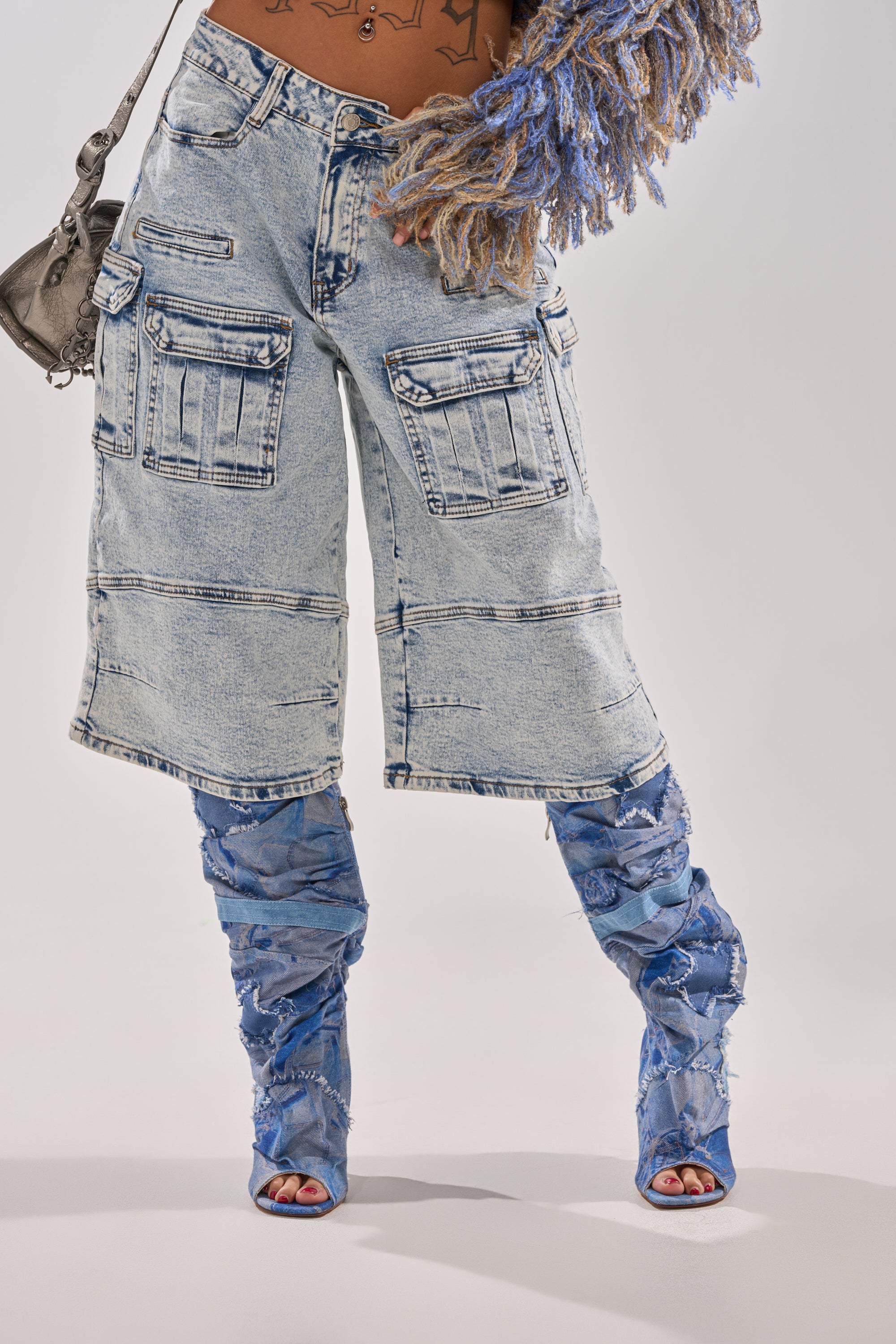 Wearing the FORGET ME NOT DENIM CARGO BERMUDA SHORTS, paired with distressed blue denim boots, a gray purse, a fringed sweater, and revealing glimpses of tattoos for an edgy look. 2