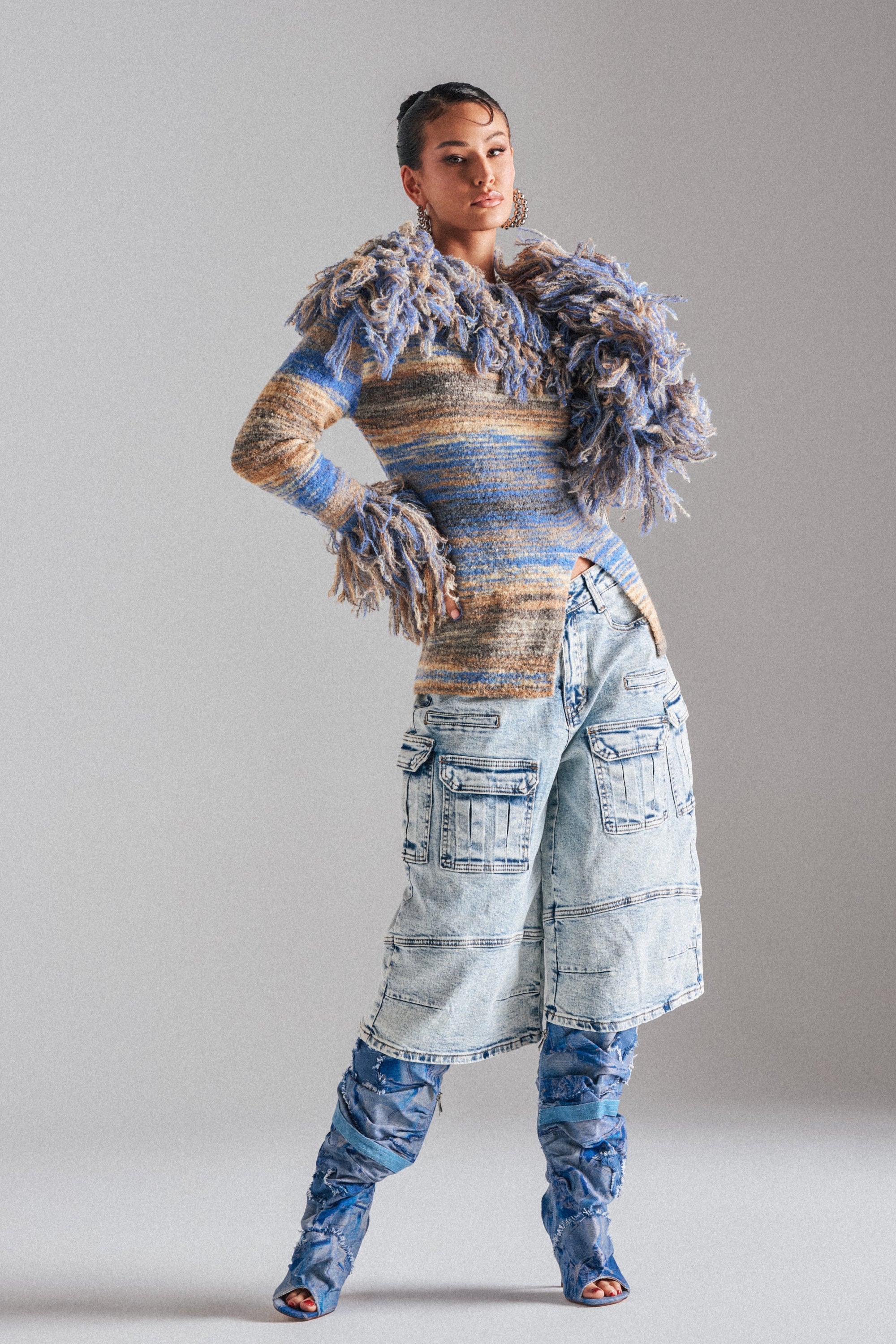 A woman poses in the FORGET ME NOT DENIM CARGO BERMUDA SHORT, paired with a multicolored textured sweater and blue knee-high boots against a plain gray background. 3