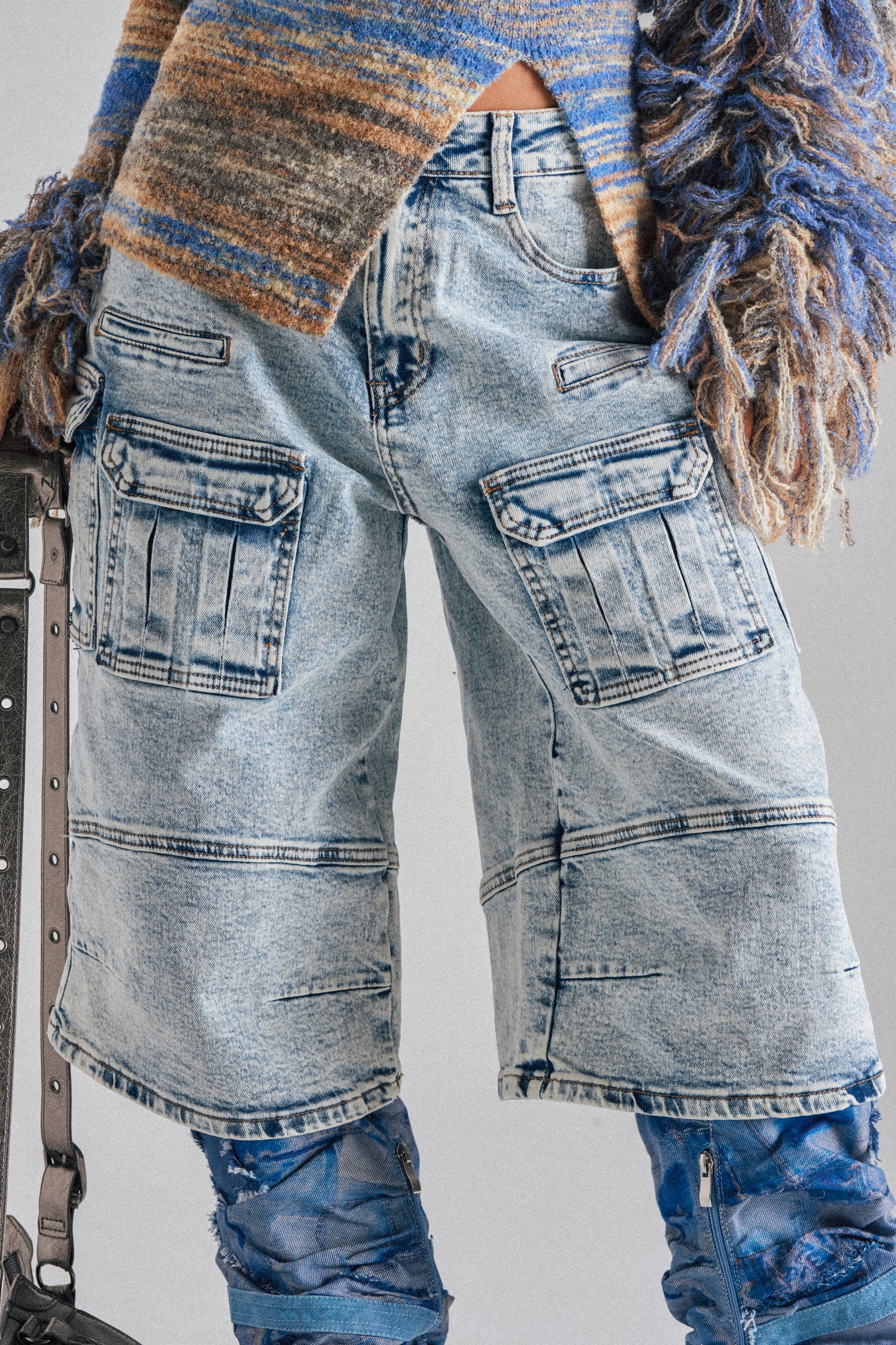 A person wearing the FORGET ME NOT DENIM CARGO BERMUDA SHORT in light blue acid-wash with multiple cargo pockets, a textured striped sweater with frayed sleeves, and blue distressed pants layered underneath. 4
