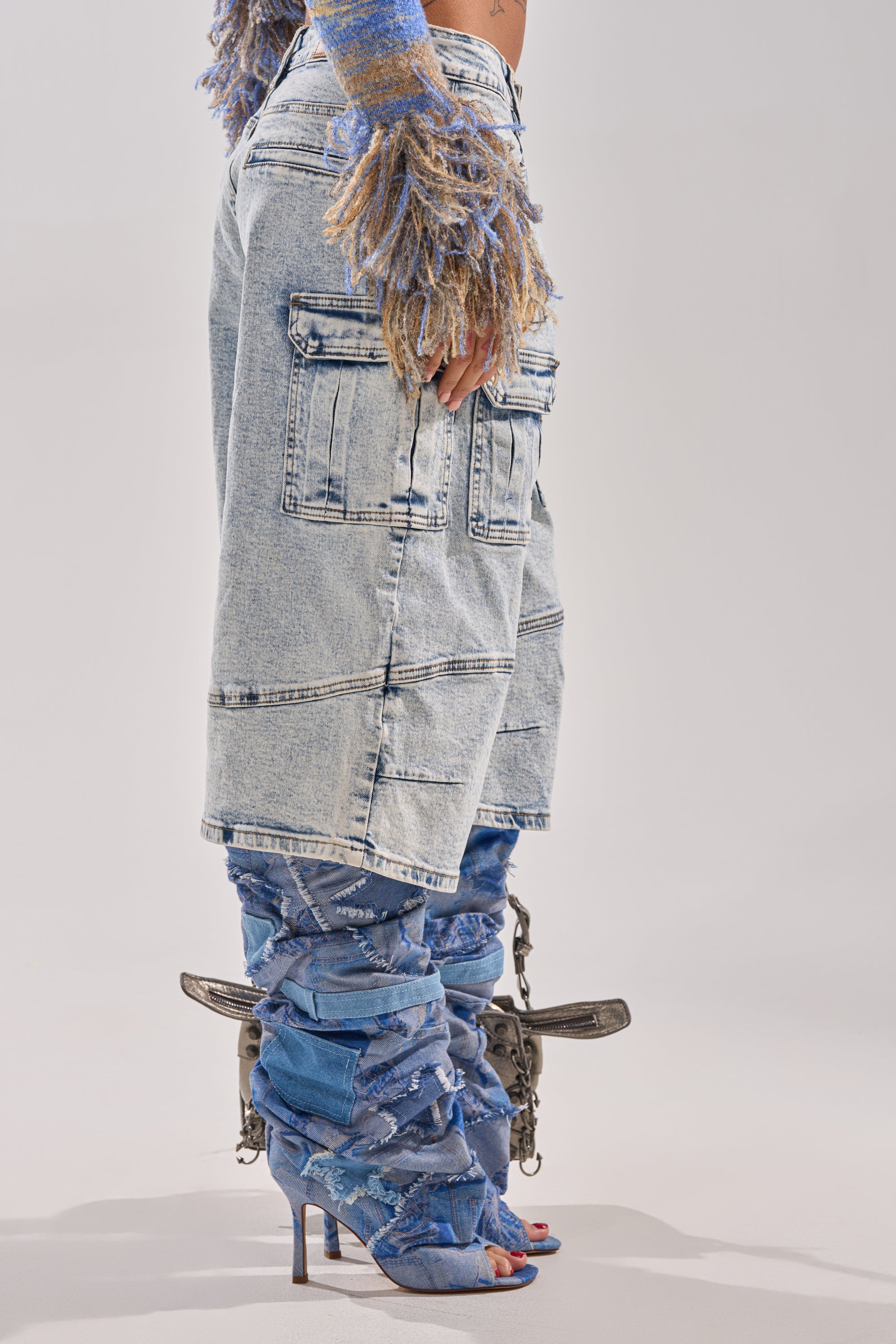A person stands sideways wearing the FORGET ME NOT DENIM CARGO BERMUDA SHORT with cargo pockets, paired with a fuzzy multicolored sweater and tall blue heeled boots that have a distressed look and metallic hardware at the heels. 7