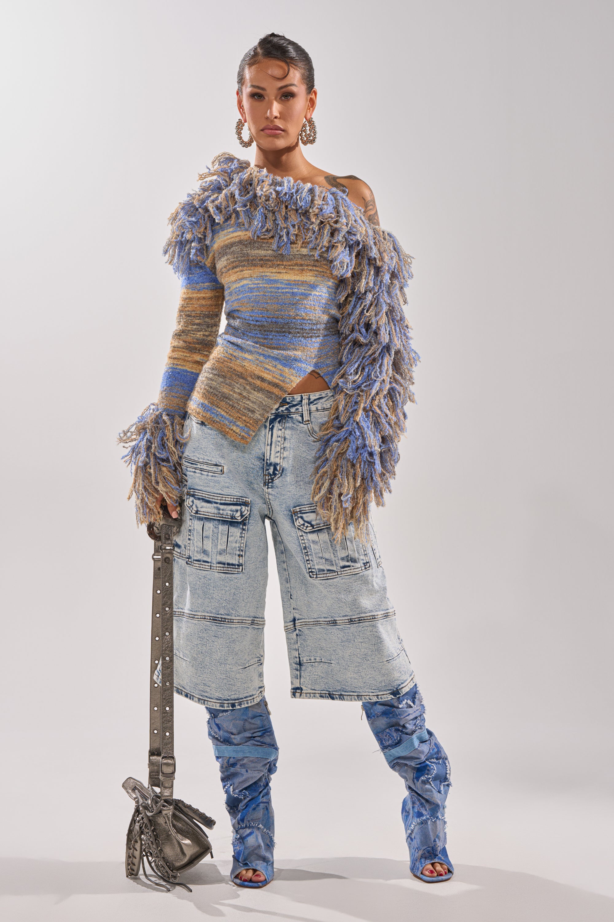 A woman stands confidently in textured, multicolored off-shoulder sweater, hoop earrings, matching denim boots, holding a silver handbag, and wearing the FORGET ME NOT DENIM CARGO BERMUDA SHORT. 9