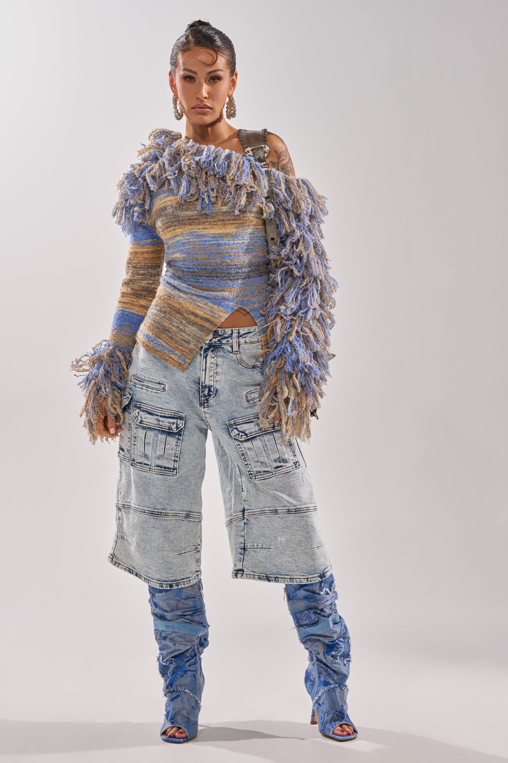 Woman stands confidently in a textured, multicolored fringe sweater, FORGET ME NOT DENIM CARGO BERMUDA SHORT with multiple pockets, and blue slouchy open-toe boots. Plain light background. 10
