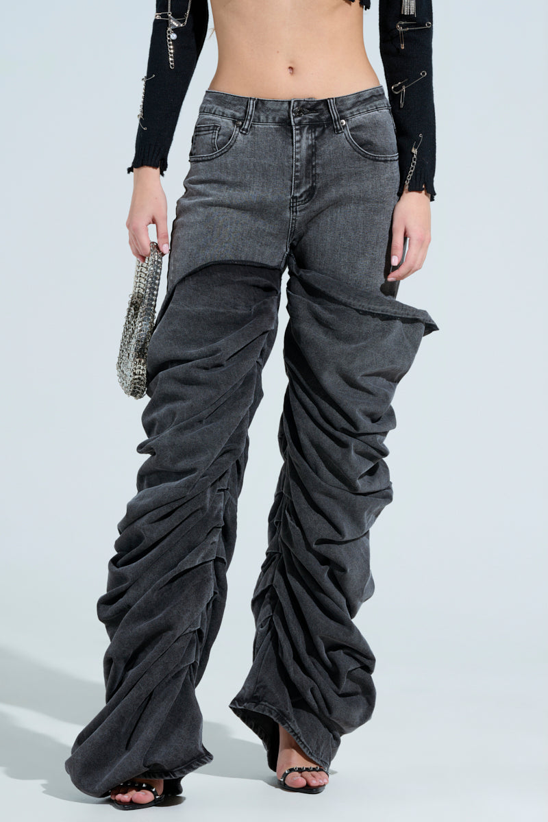 Wearing the "OH YEAH BABY BLACK WASH DENIM PANT," a person in relaxed mid-rise black denim stands against a plain background, pairing them with a black cropped top and accessorizing with a small silver handbag and open-toed black heels.