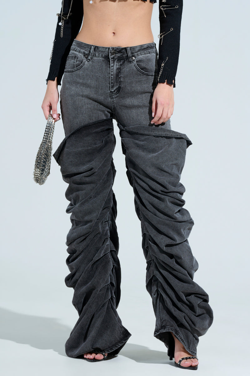 Wearing the OH YEAH BABY BLACK WASH DENIM PANT with a relaxed mid-rise fit, they pair it with a cropped black top accented by metal pins. A small silver chain bag complements high-heeled sandals, and the plain background highlights this chic ensemble perfectly.