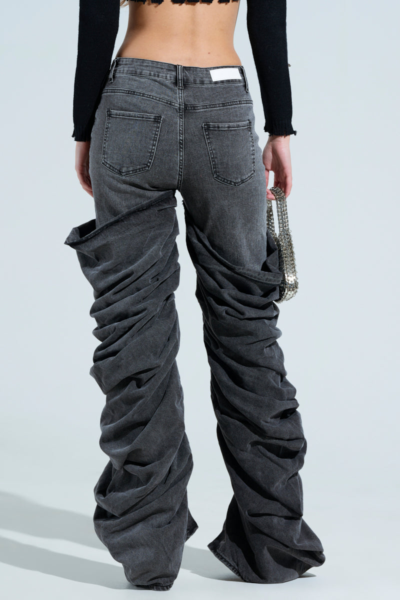 A person wears oversized OH YEAH BABY black wash denim pants with a relaxed mid-rise fit and pairs them with a cropped black top, holding a small metallic handbag. They are seen from behind against a neutral background.