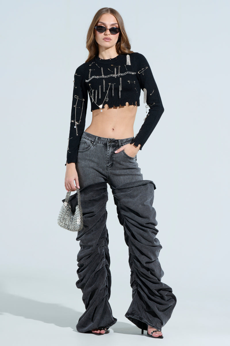 Wearing sunglasses, a person confidently poses in a black cropped top with metallic accents and OH YEAH BABY BLACK WASH DENIM PANT, offering a relaxed mid-rise fit. They hold a textured handbag against a light blue background.