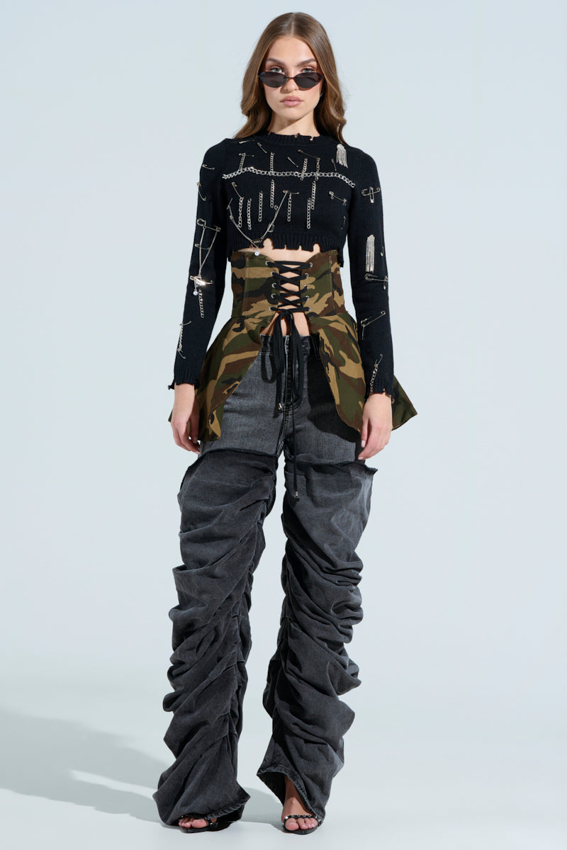 A woman in sunglasses wears a black top with silver pins, a camouflage corset, and OH YEAH BABY BLACK WASH DENIM PANT oversized ruched tonal pants in a relaxed mid-rise fit. She stands against a light blue background.