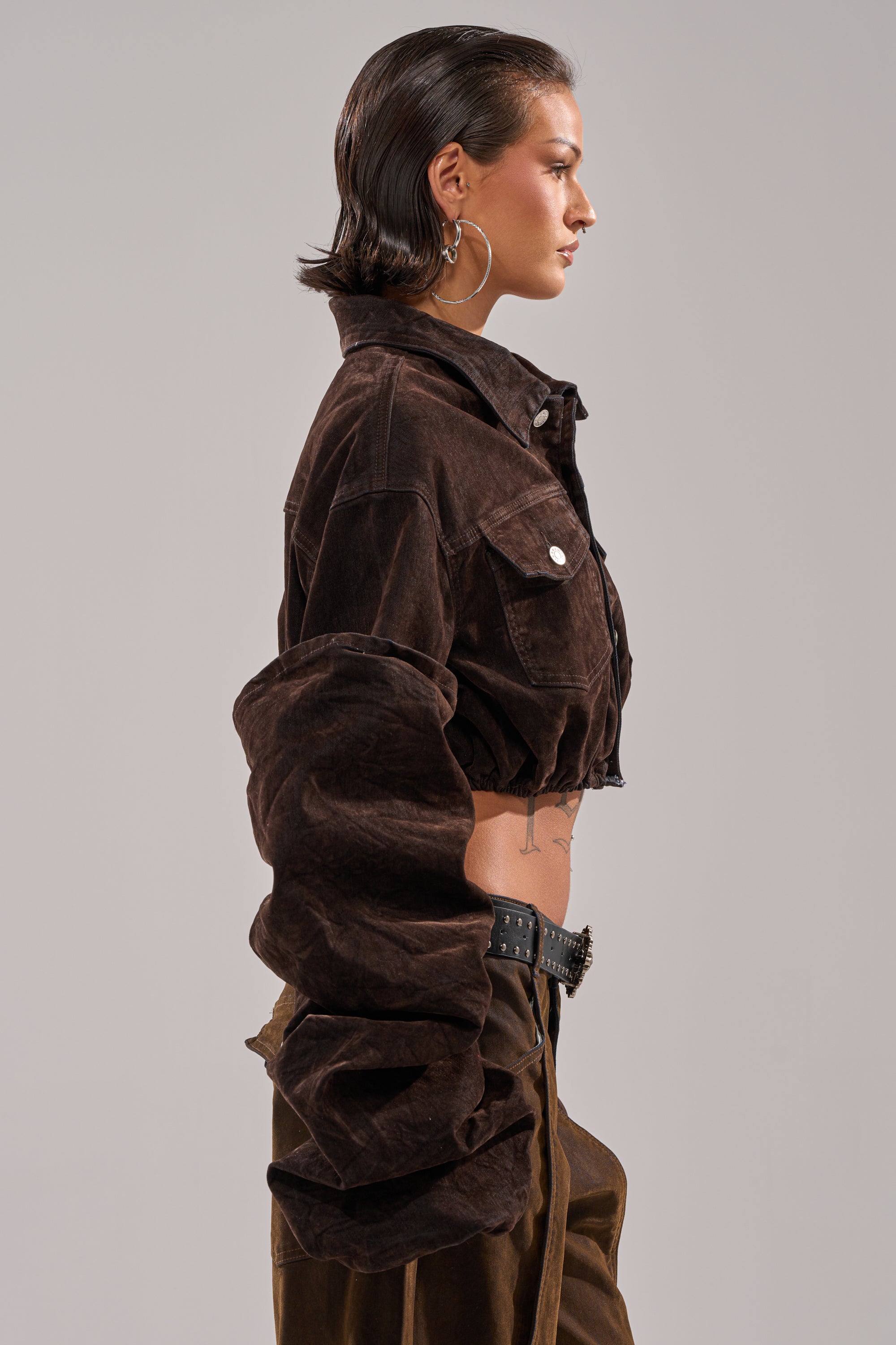 A person with short slicked-back hair wears the STACKED SLEEVE VELVET DENIM JACKET and matching pants, paired with a black studded belt and large hoop earrings, standing in profile against a plain light background.