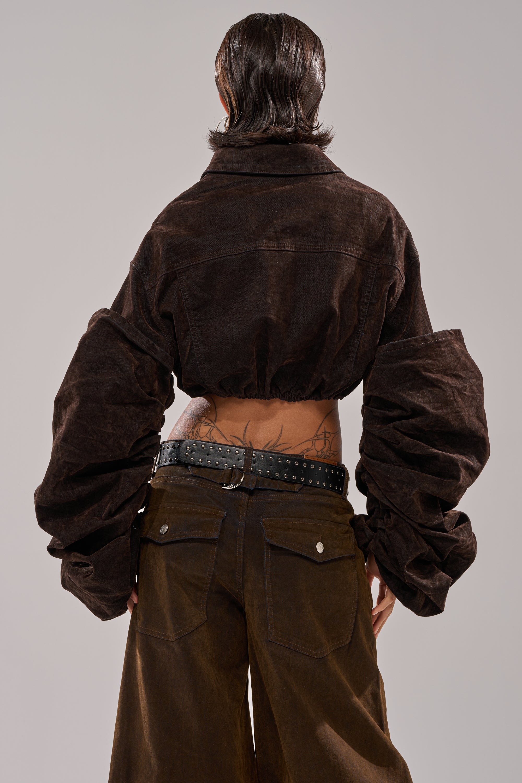 Person stands with their back to the camera, wearing the STACKED SLEEVE VELVET DENIM JACKET with oversized sleeves and wide-leg pants. Their exposed midriff features tattoos and a studded black belt.
