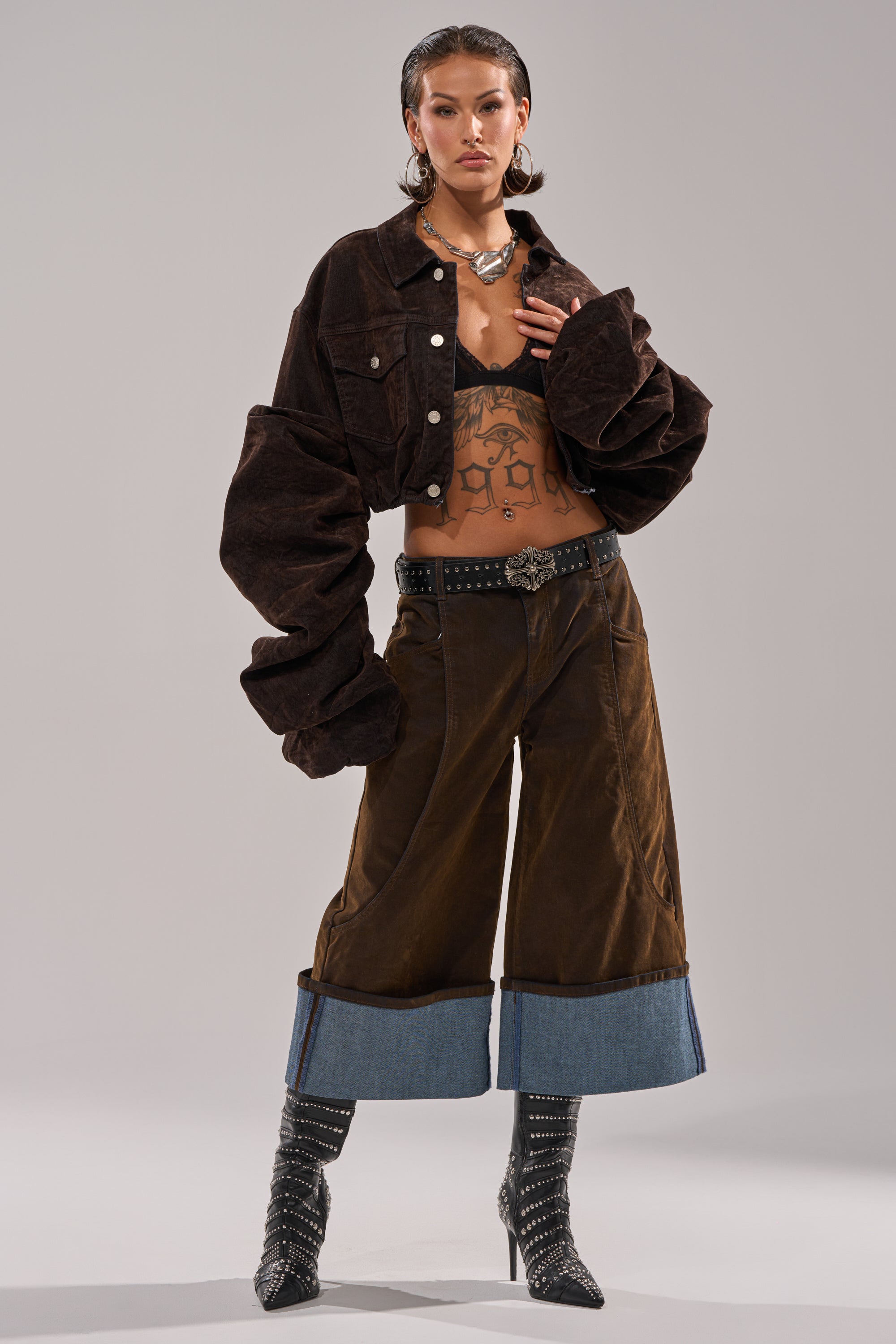 A person stands confidently in the STACKED SLEEVE VELVET DENIM JACKET, wide-legged brown pants with blue denim cuffs, a studded belt, pointed heeled boots, layered necklaces, statement earrings, and slicked-back hair.