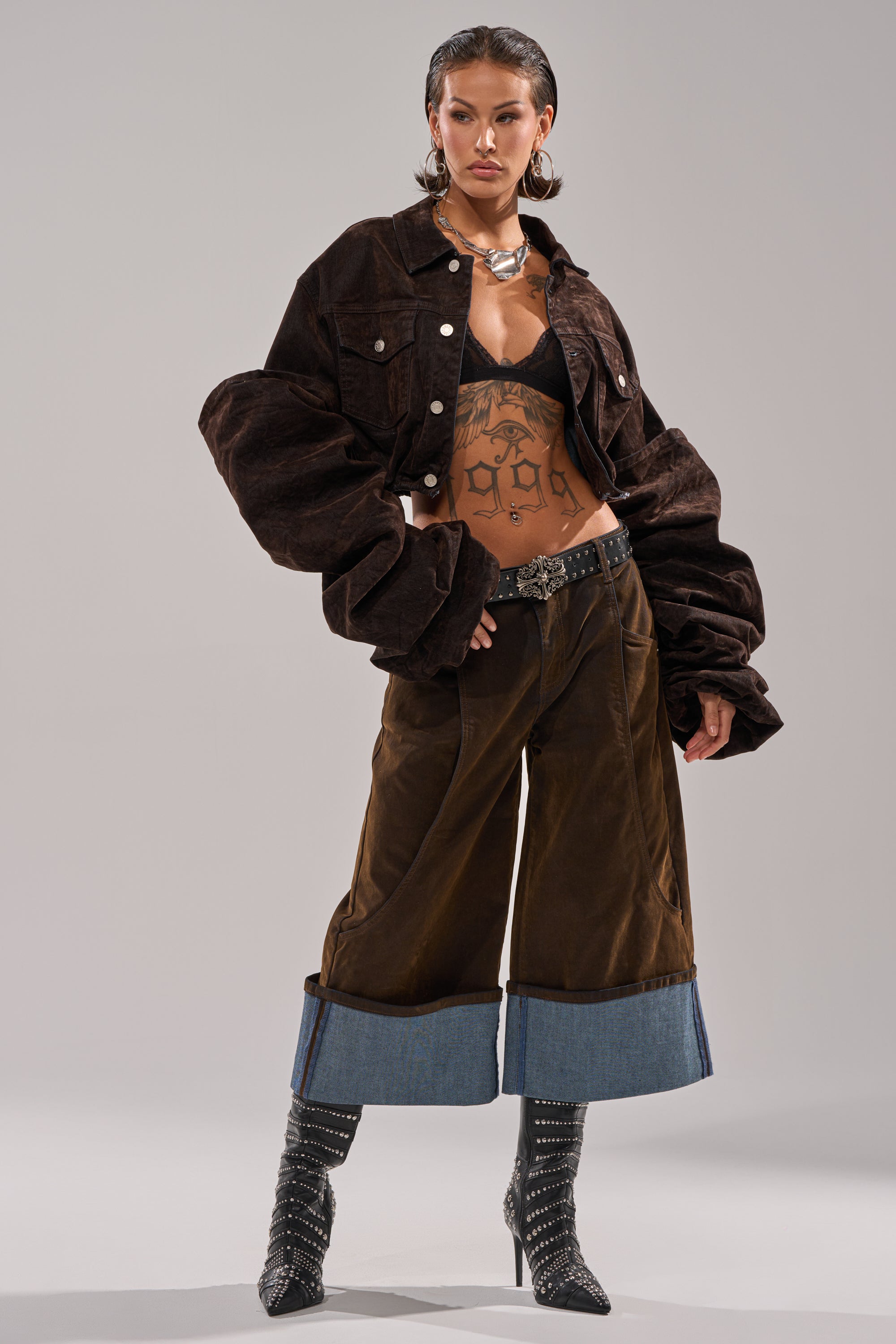 A woman poses confidently in the STACKED SLEEVE VELVET DENIM JACKET, paired with loose brown pants cuffed in denim, studded high-heeled boots, layered jewelry, and showing off her torso tattoos.