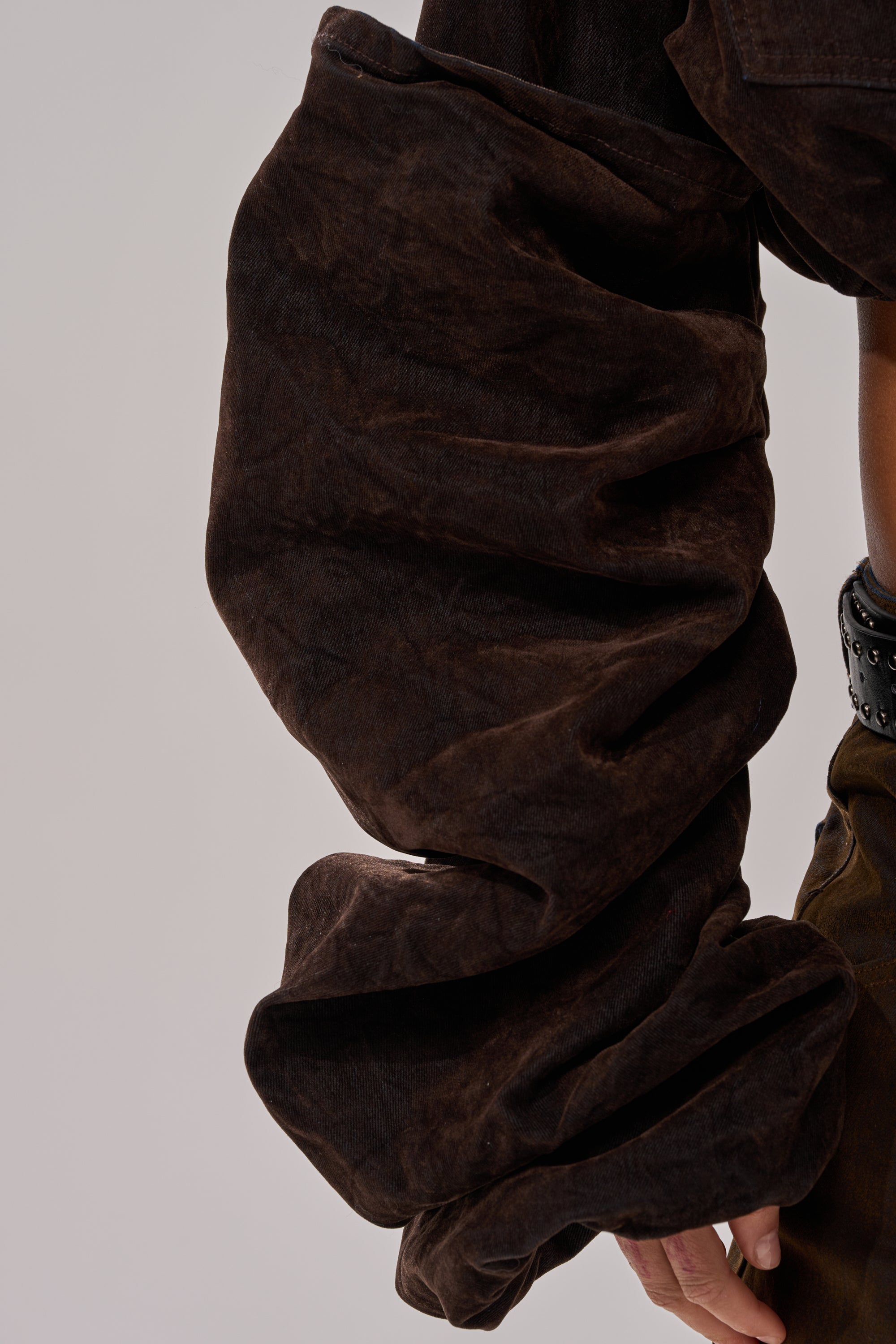 A close-up shows a person wearing the STACKED SLEEVE VELVET DENIM JACKET in brown with oversized, gathered sleeves, styled with a studded black belt. The plain, neutral background highlights the jacket's unique details.