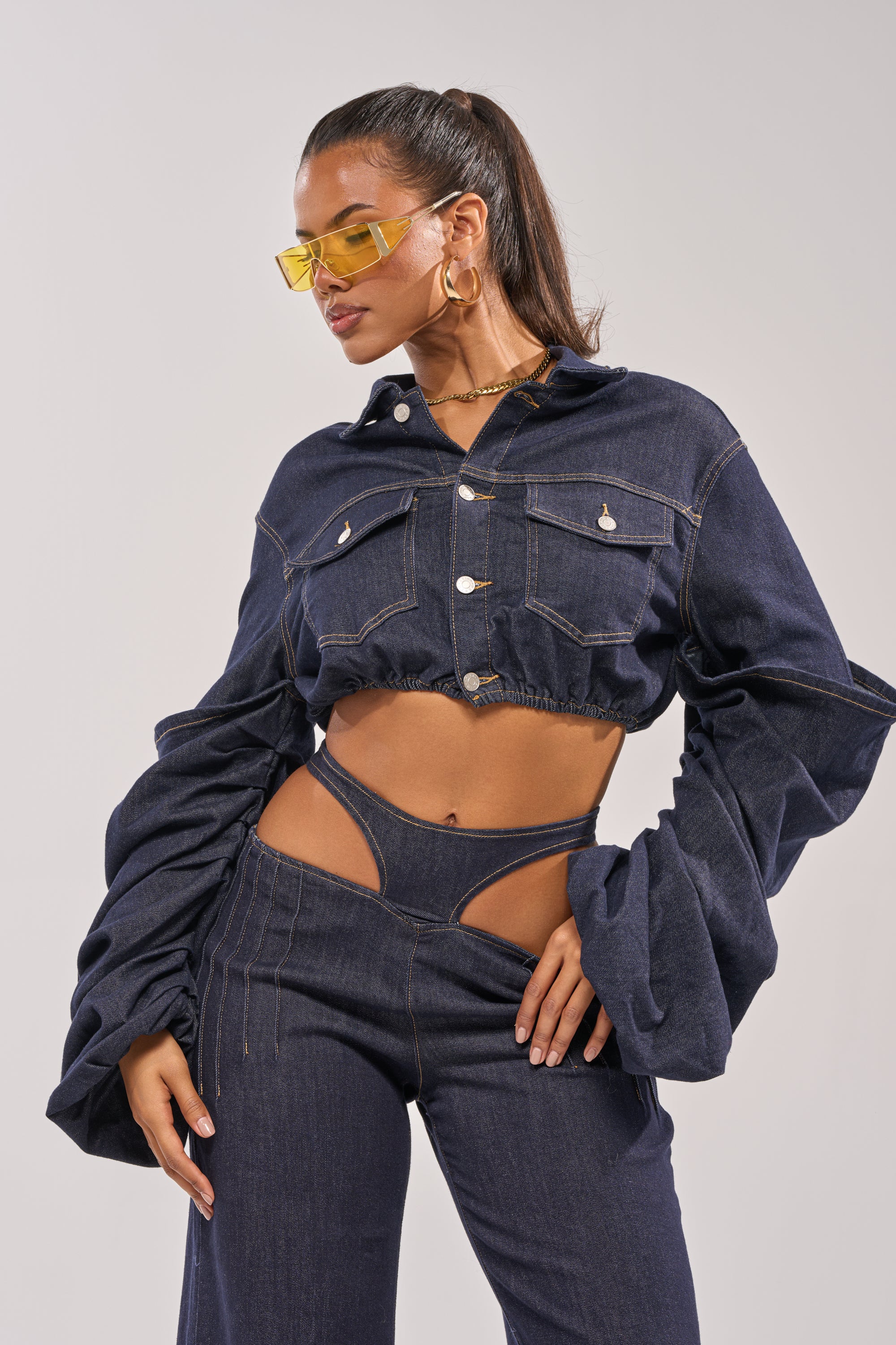 A woman poses in the STACKED SLEEVE CROPPED DENIM JACKET with oversized sleeves and high-waisted cutout pants, styled with gold hoop earrings, a chain necklace, and yellow-tinted sunglasses for a bold layered look. 0