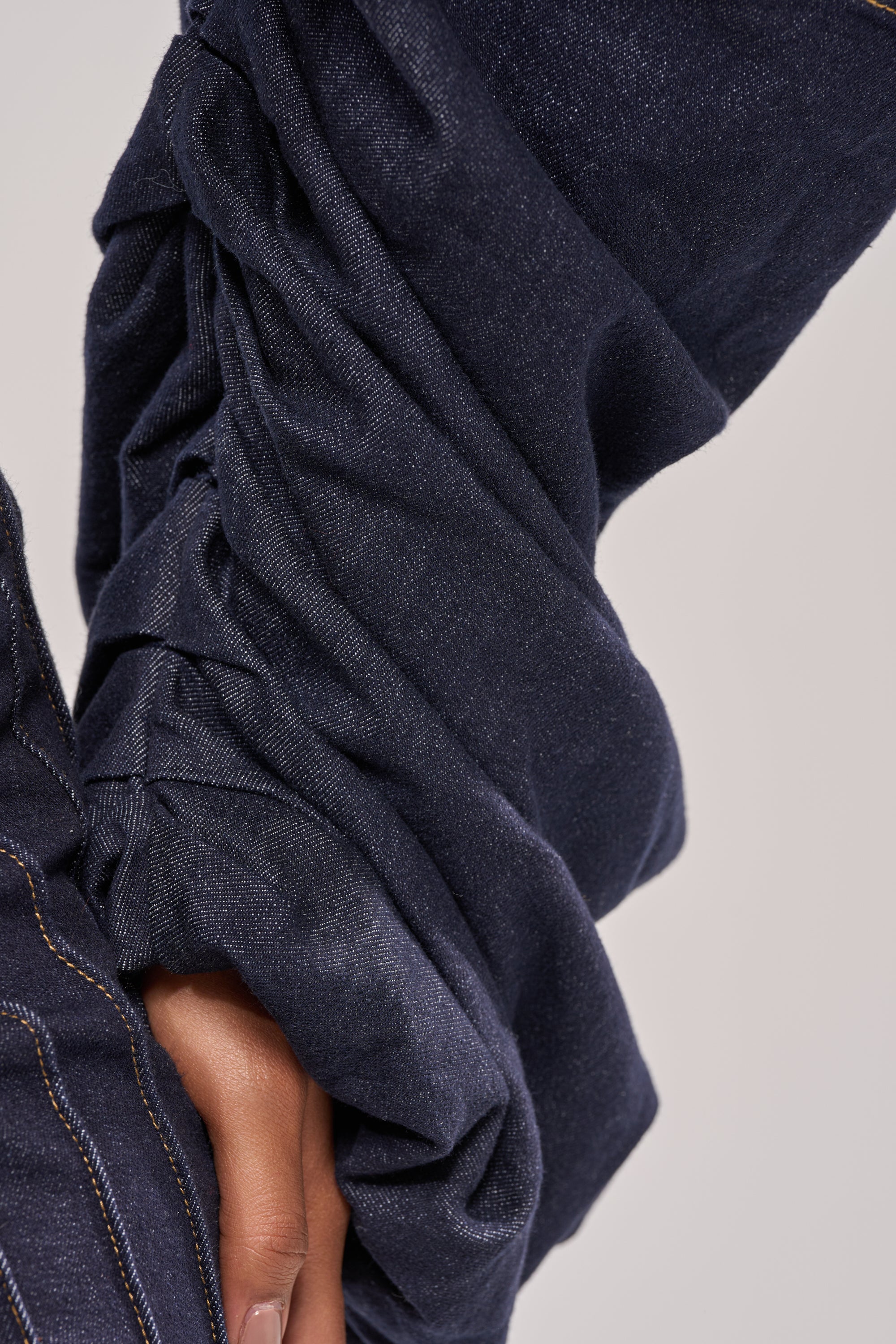 A close-up of a hand gently holding the ruched sleeve of the STACKED SLEEVE CROPPED DENIM JACKET highlights its visible stitching details against a soft, neutral backdrop. 10
