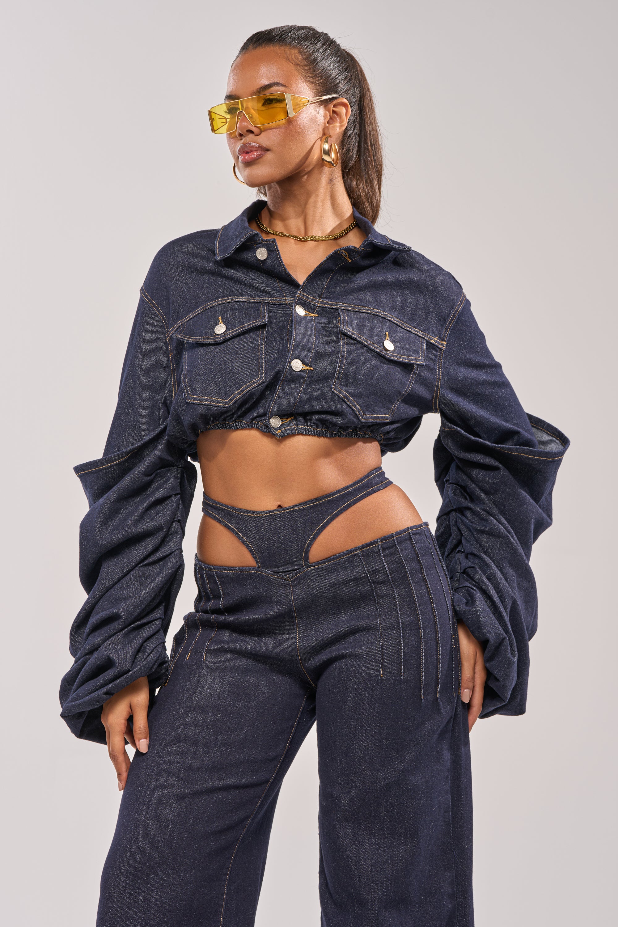 A woman models the STACKED SLEEVE CROPPED DENIM JACKET, styled with high-waisted cut-out pants, yellow-tinted sunglasses, and gold hoop earrings for a bold look. 3