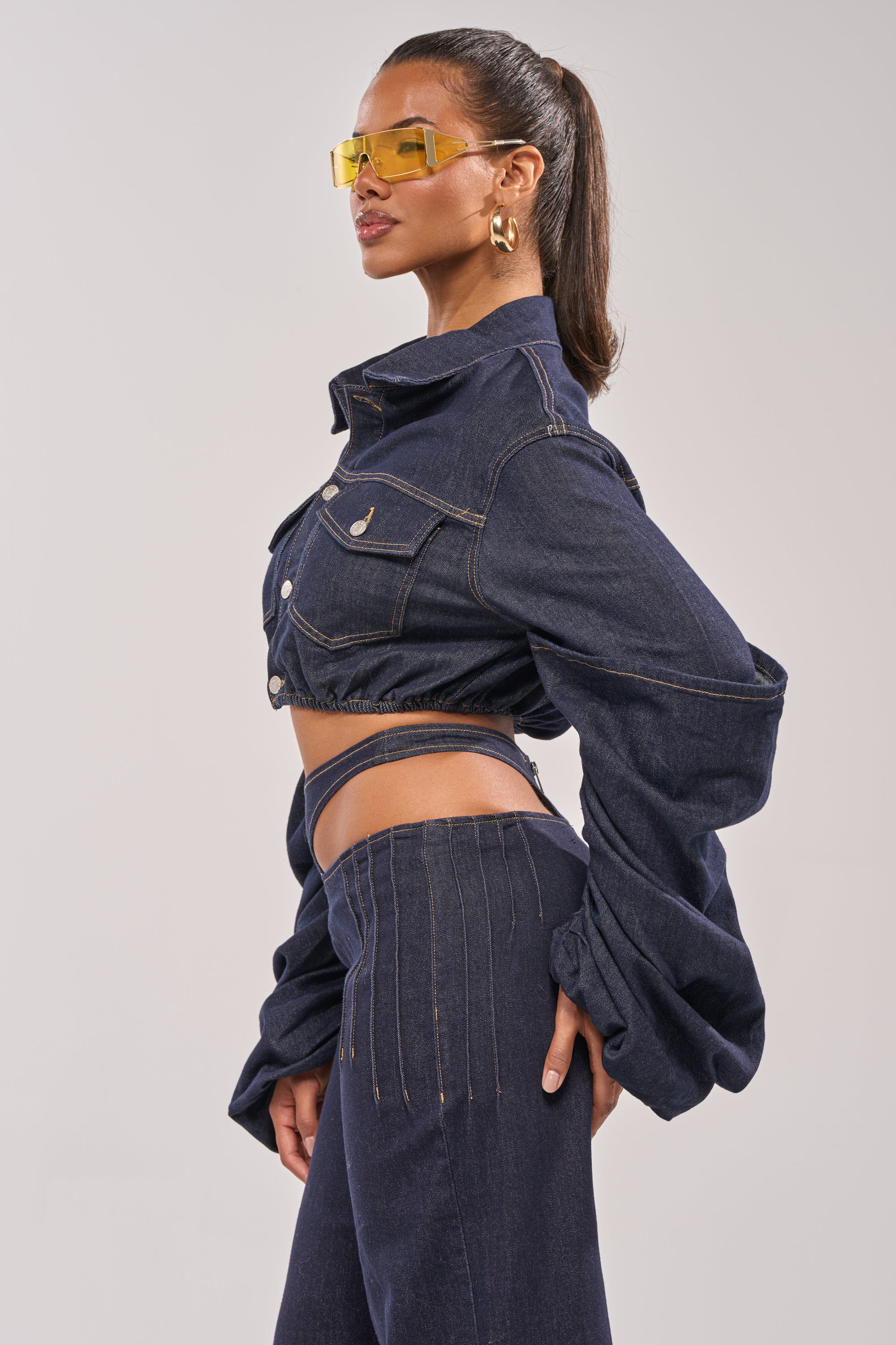 A woman models the STACKED SLEEVE CROPPED DENIM JACKET with high-waisted jeans, styled with gold hoops, yellow sunglasses, and a sleek ponytail against a plain background for a bold layered look. 5