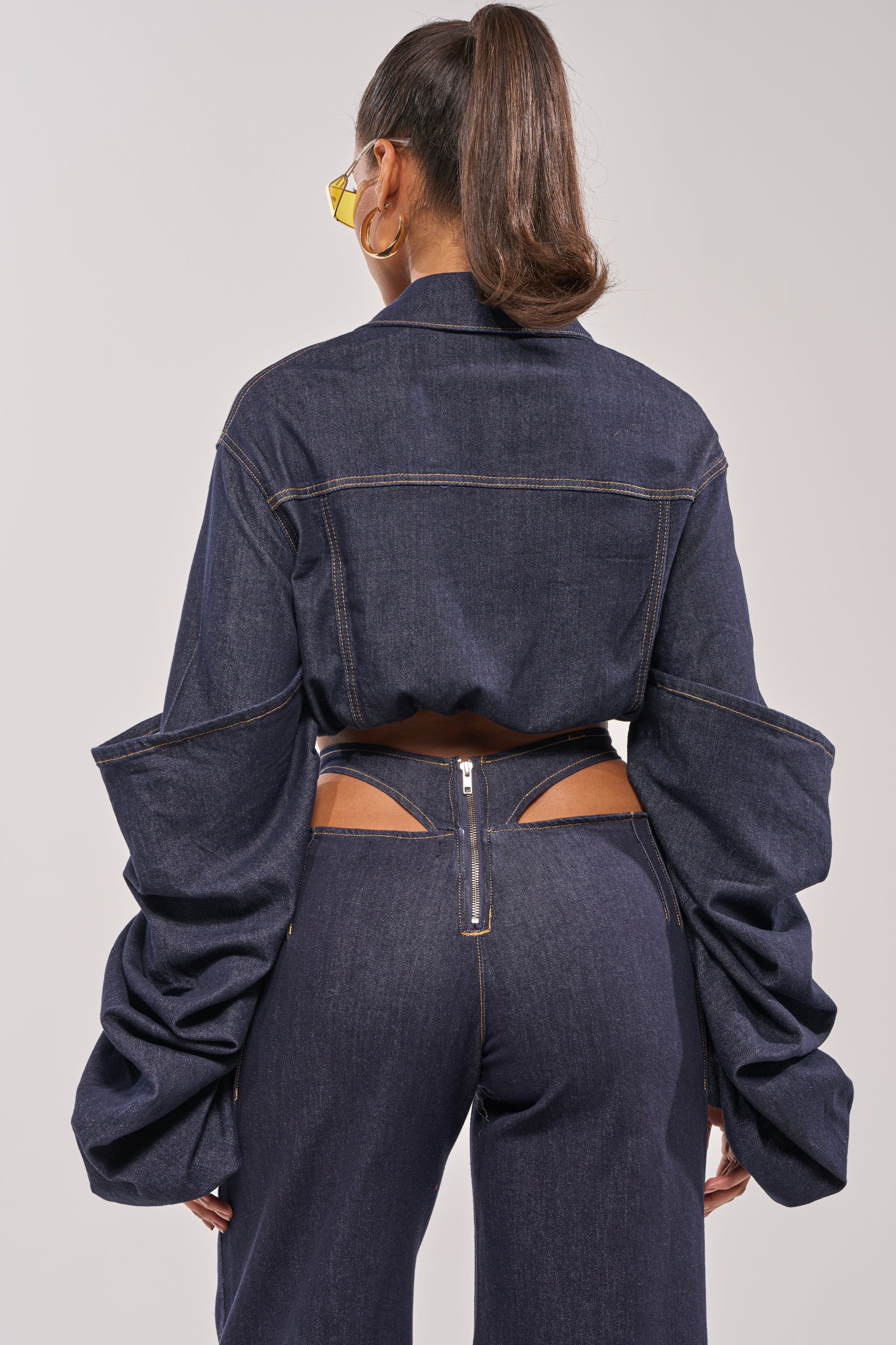 A person faces away, wearing dark blue denim pants with a back cutout and zipper, paired with the STACKED SLEEVE CROPPED DENIM JACKET featuring sculptural, exaggerated sleeves. They have a high ponytail, yellow sunglasses, and hoop earrings. 6