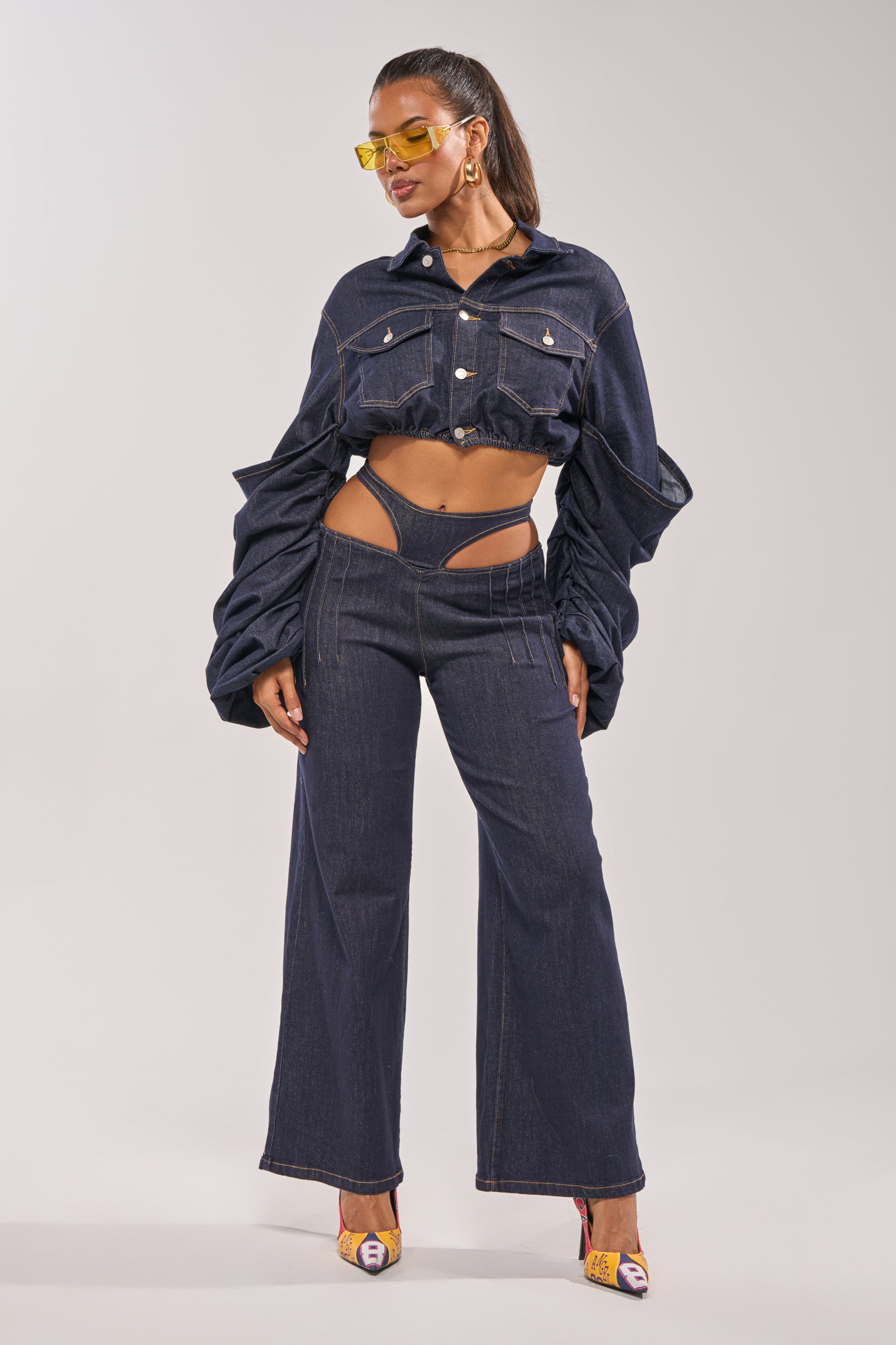 A woman poses in the STACKED SLEEVE CROPPED DENIM JACKET, featuring sculptural, exaggerated sleeves. She pairs it with high-waisted, wide-leg jeans with hip cut-outs, yellow sunglasses, and patterned heels for a bold layered look. 7