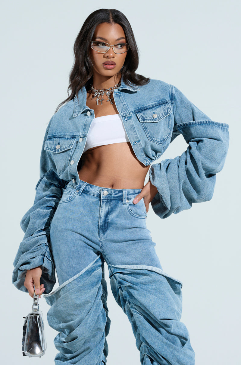 A person in a STACKED SLEEVE DENIM JACKET and ruched jeans wears a white cropped top. Glasses glint behind a silver necklace as they hold a metallic handbag against a plain background.