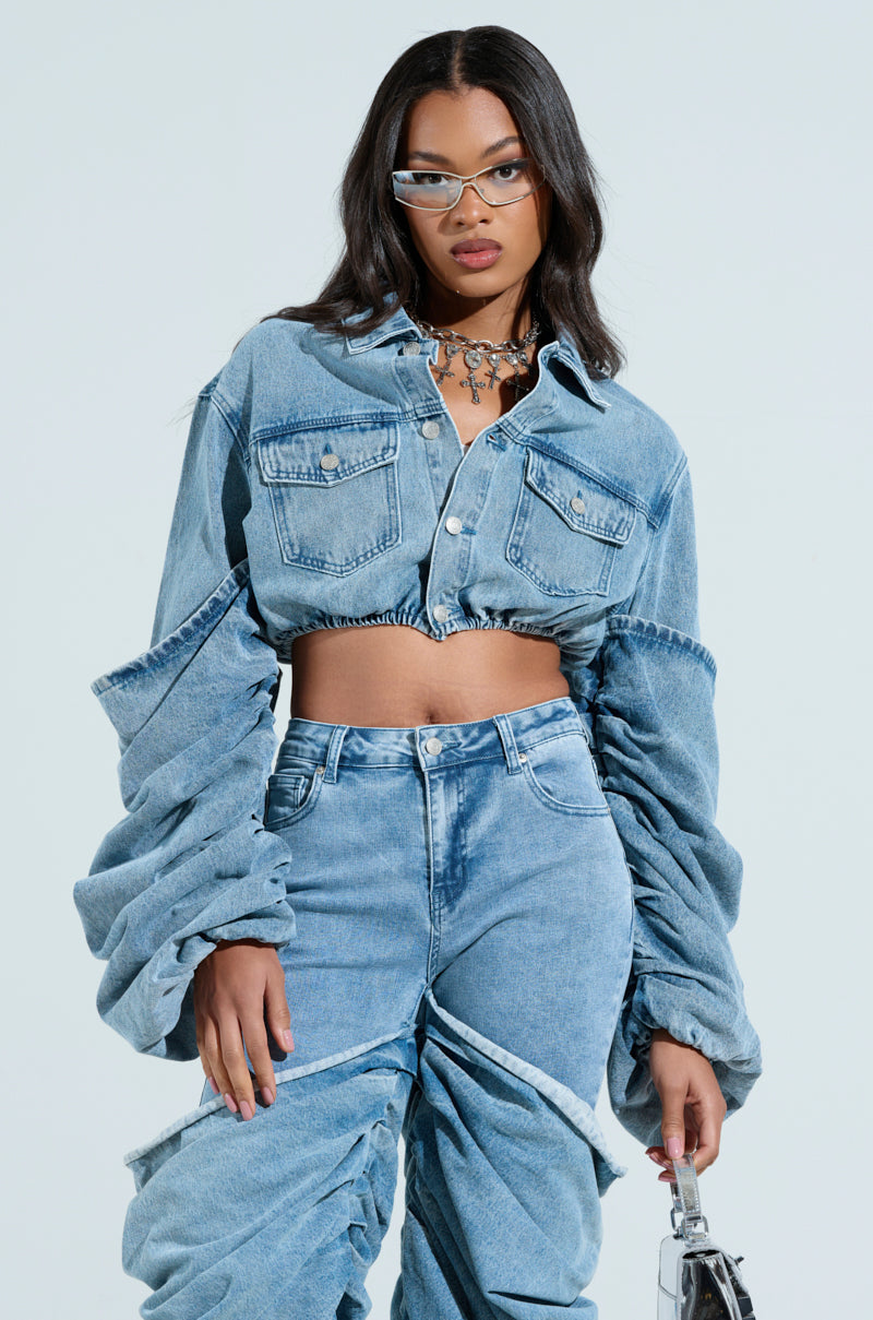 A woman confidently poses in a STACKED SLEEVE DENIM JACKET and matching jeans, accessorized with clear glasses, a necklace, and carrying a small bag against a plain background.