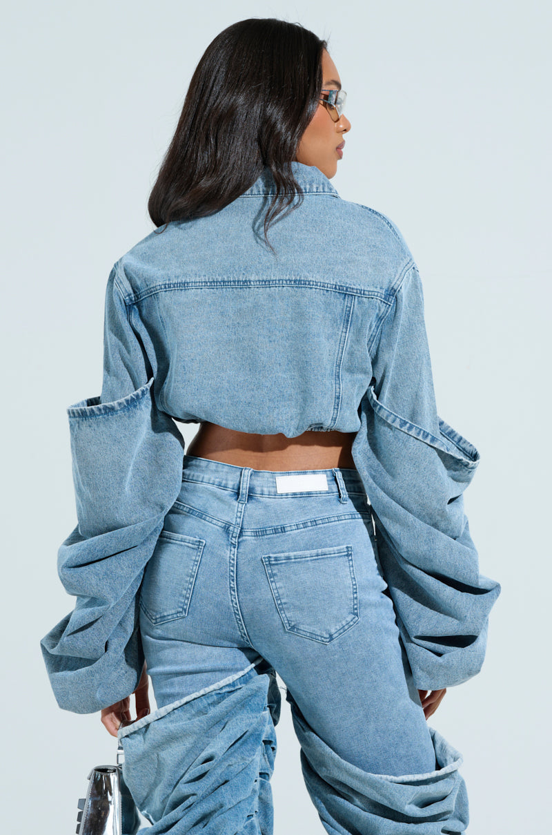 A person with long hair, wearing a STACKED SLEEVE DENIM JACKET with stylish oversized elements and ruched denim pants, stands back turned. They hold a transparent bag and wear sunglasses. The cropped fit adds to the edgy design.